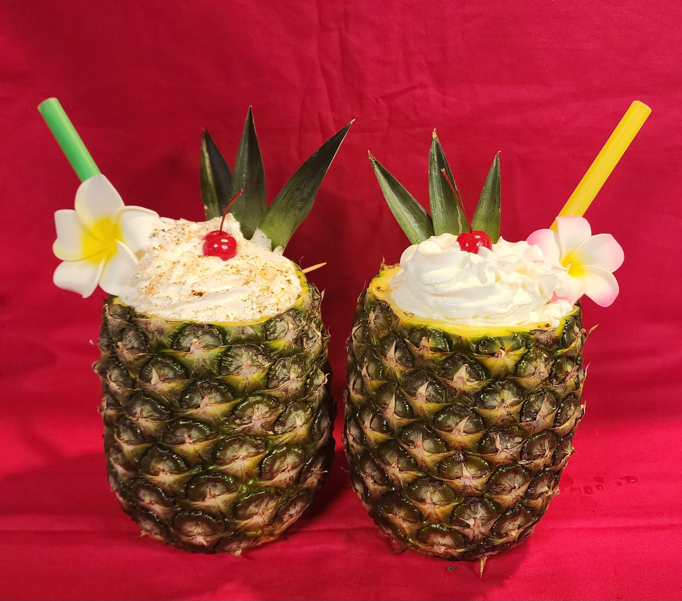 Two hollowed-out pineapples filled with whipped cream, garnished with cherries, pineapple leaves, and flowers, with colorful straws, on a red background.