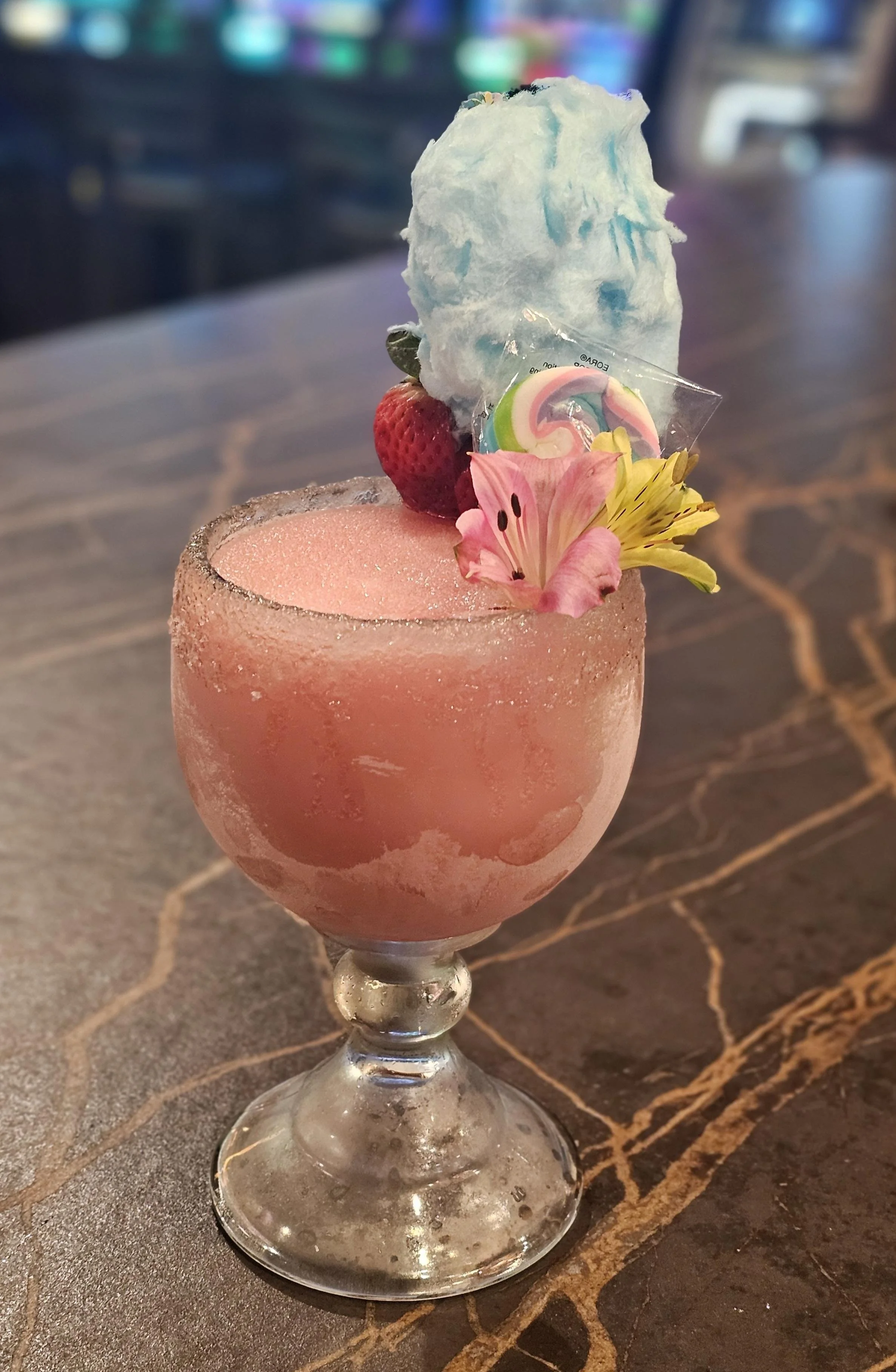 A pink frozen cocktail garnished with a strawberry, flowers, rainbow lollipop, and a large dollop of cotton candy in a stemmed glass.