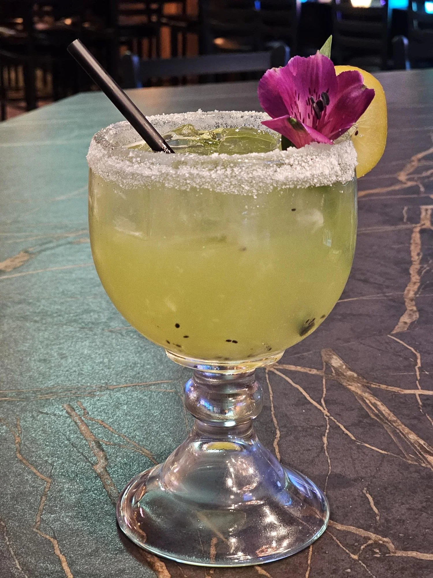 A yellow cocktail in a large glass goblet, rimmed with sugar, garnished with a pink flower and lemon slice, with a black straw inside, placed on a marble table.