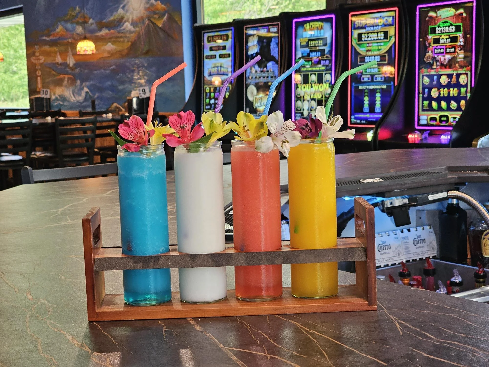 Four colorful drinks garnished with flowers and straws are placed on a table in a bar or casino with slot machines in the background.