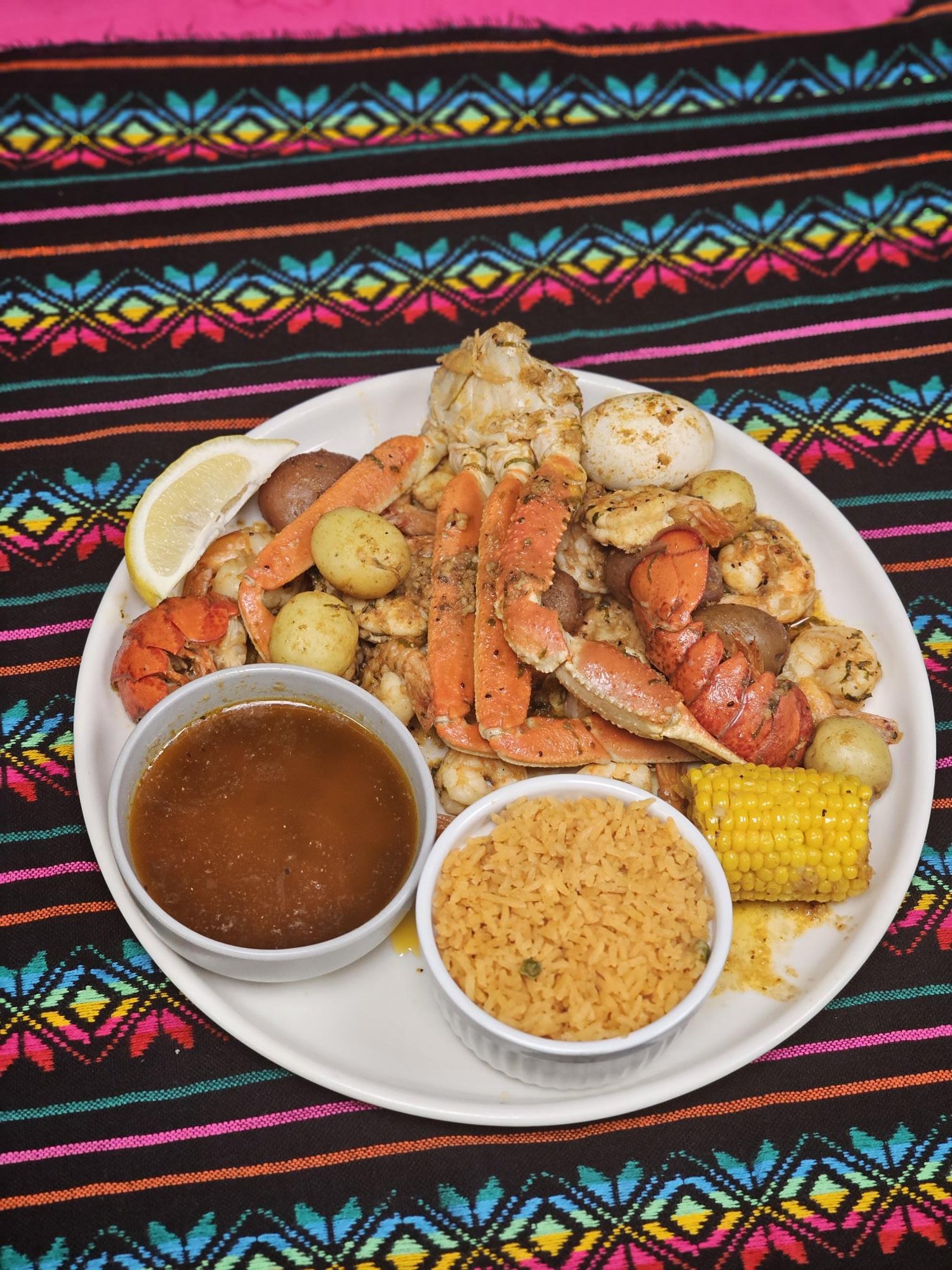 Plate of seafood, including crab legs, shrimp, boiled eggs, and baby potatoes, served with lemon and broth on a decorative colorful woven tablecloth.