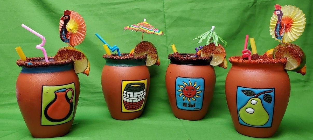 Four terracotta mugs decorated with colorful stickers of a pitcher, a grill, a sun with a face labeled 'El Sol', and a pear with leaves, each with a character-themed straw, lime wedge, and paper umbrella, set against a green background.
