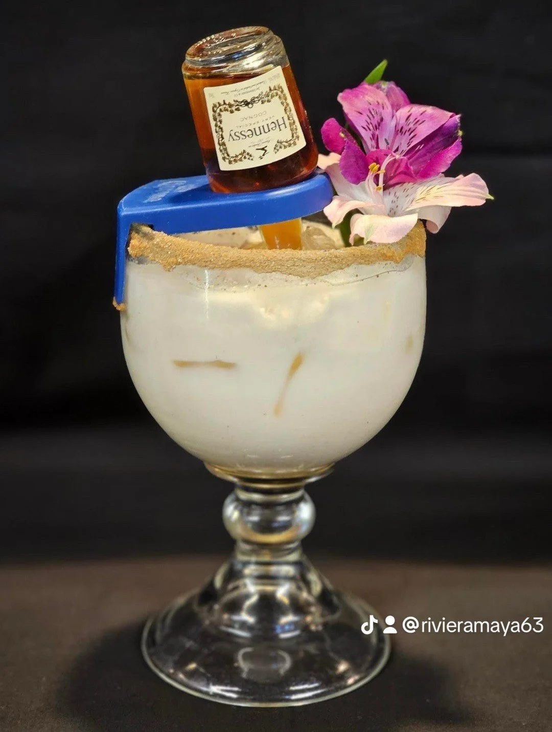 A glass dessert cup filled with a creamy dessert topped with edible flowers, a sugar rim, a blue and white chocolate garnish, a small jar of honey, and a pink flower.