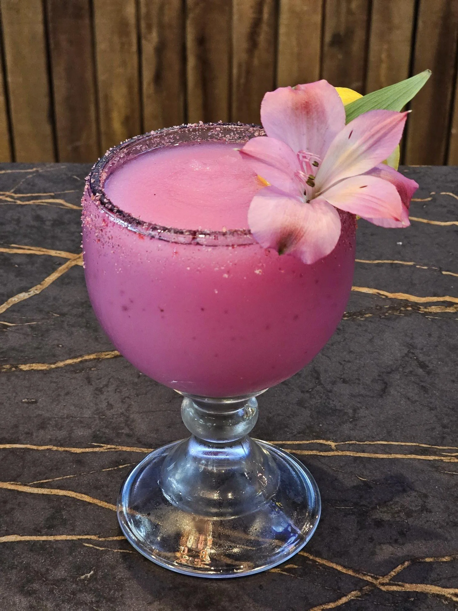 Pink cocktail in a stemmed glass garnished with pink flowers, placed on a dark surface with a wooden background.