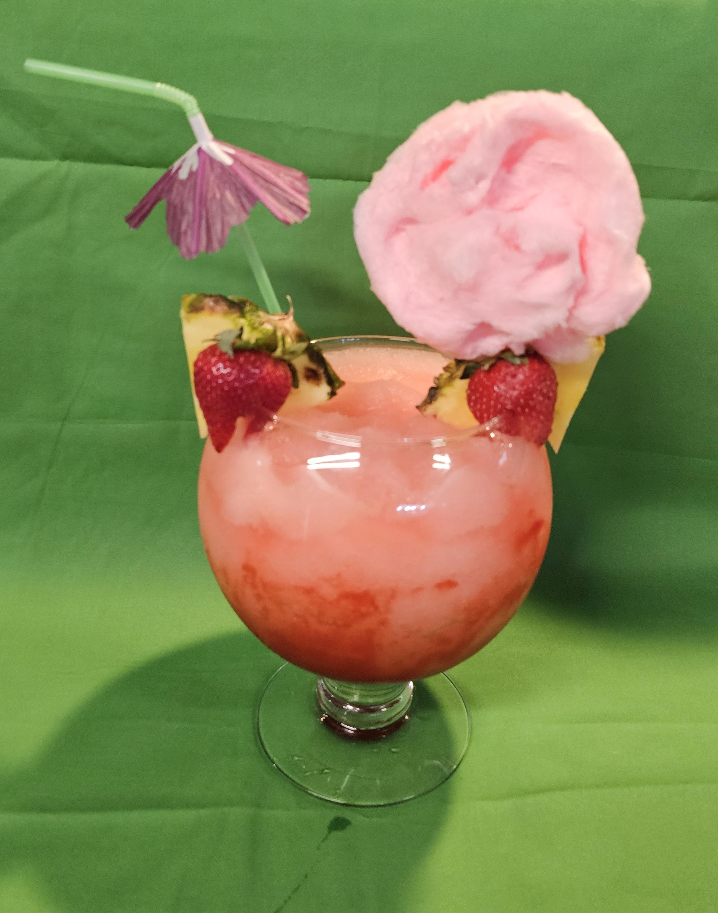 A tropical cocktail with pinkish-orange swirls, garnished with strawberries, pineapple, cotton candy, a paper umbrella, and a straw on a green table.
