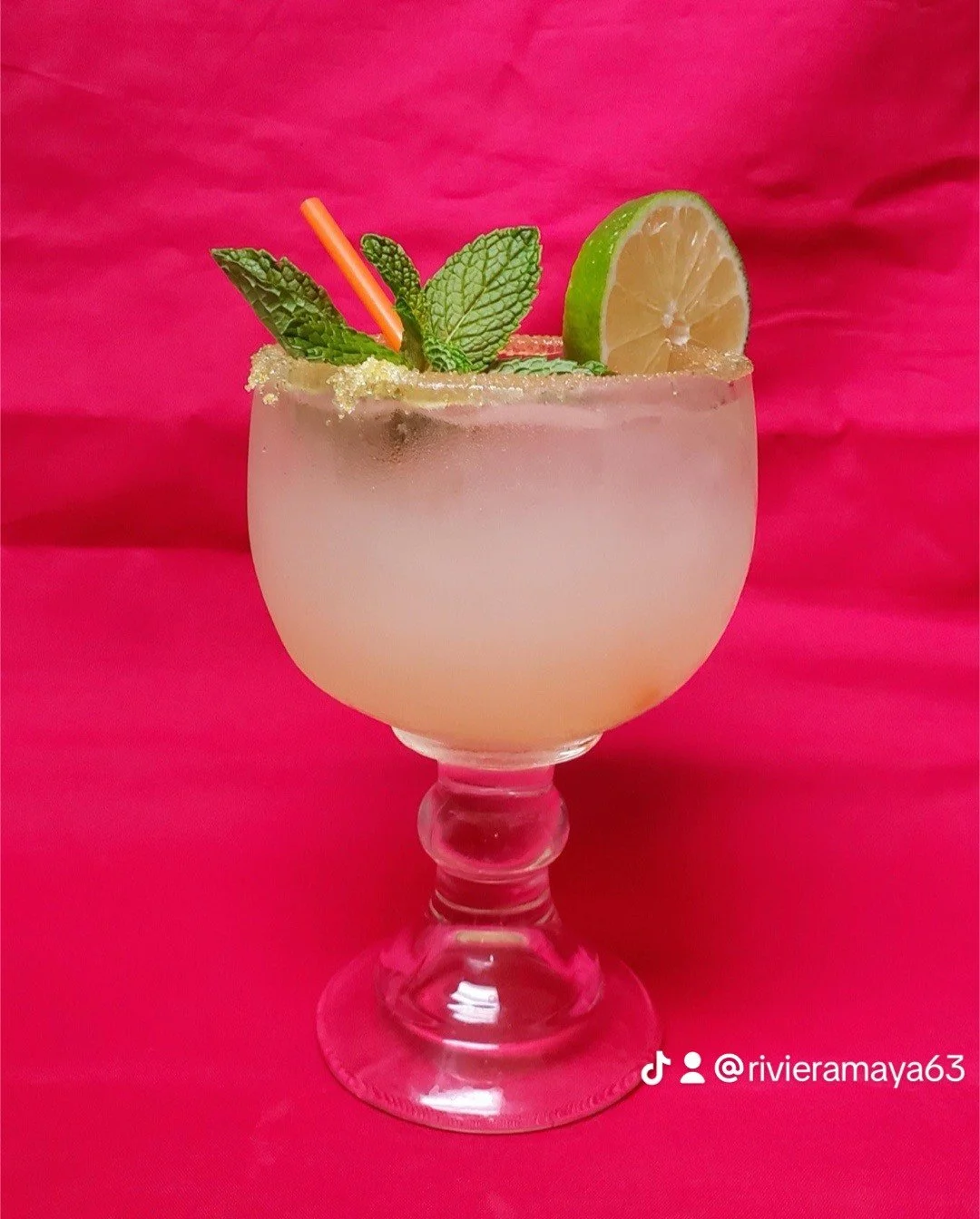 A pale pink cocktail in a large goblet glass garnished with a lime wedge, fresh mint leaves, and a pink straw, set against a pink background.
