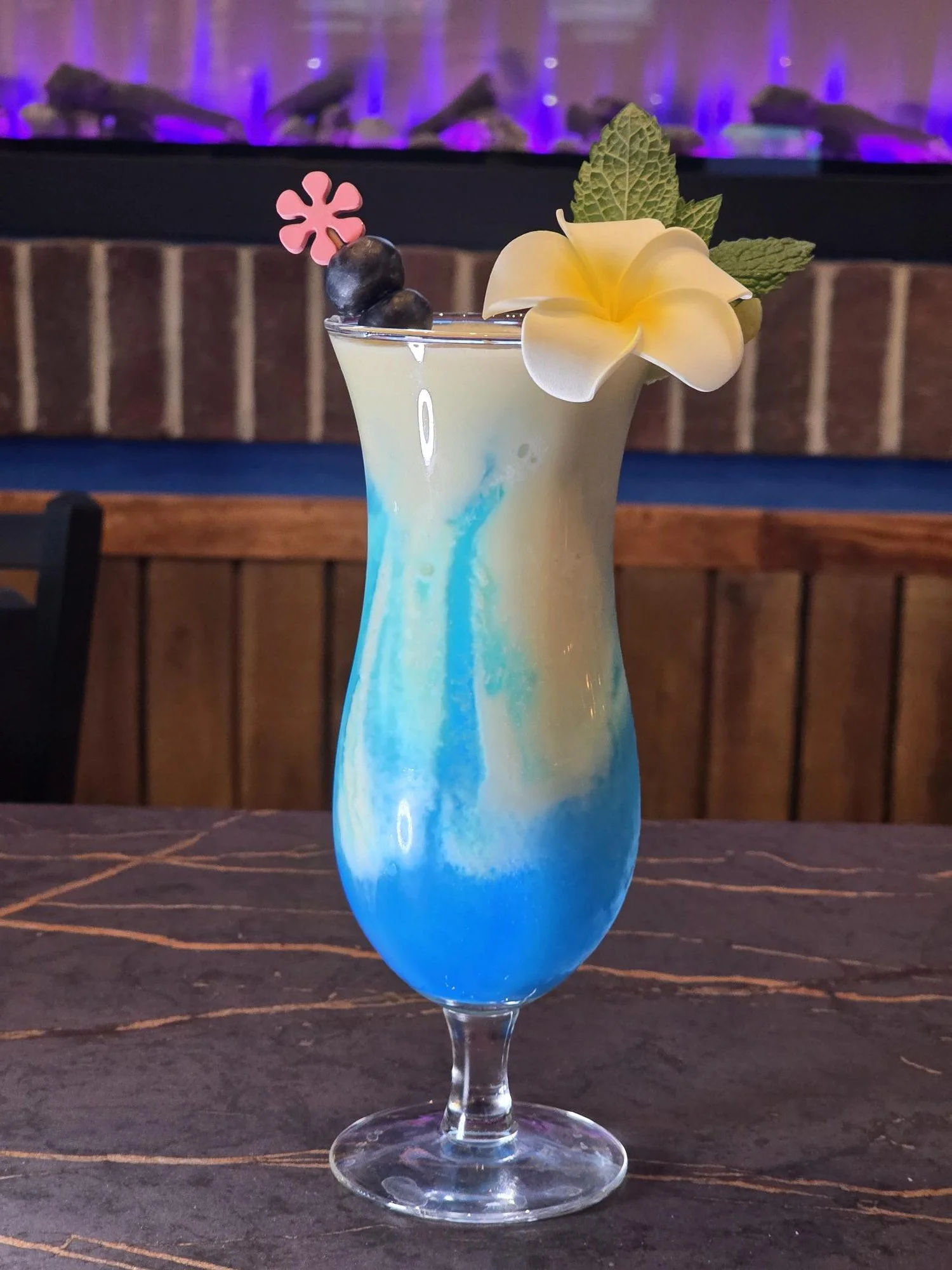 Colorful layered cocktail in a tall glass garnished with blueberries, a pink flower-shaped pick, a white flower, and mint leaves. In the background, there is a brick wall and a dark-colored surface.