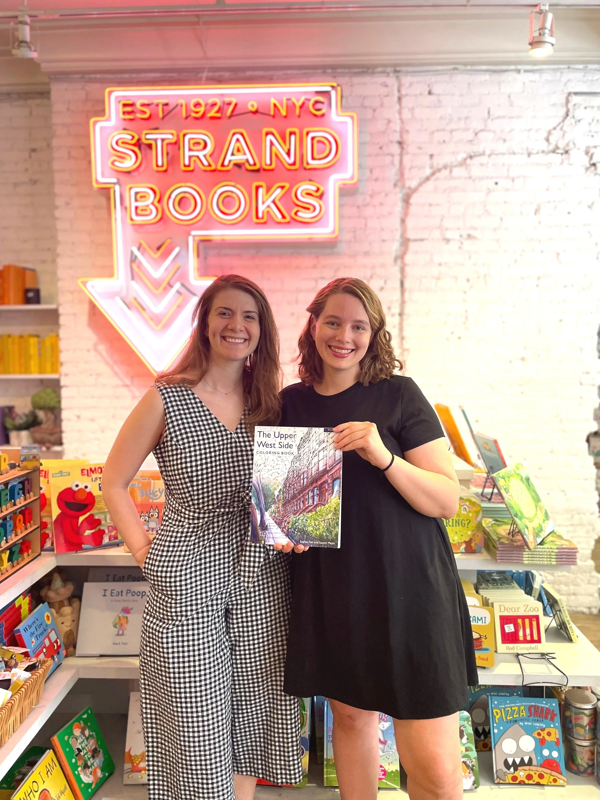Two smiling women standing in a bookstore in front of a neon sign that reads 'Strand Books' with an arrow pointing downward. One woman is holding a book titled 'The Upper West Side'.