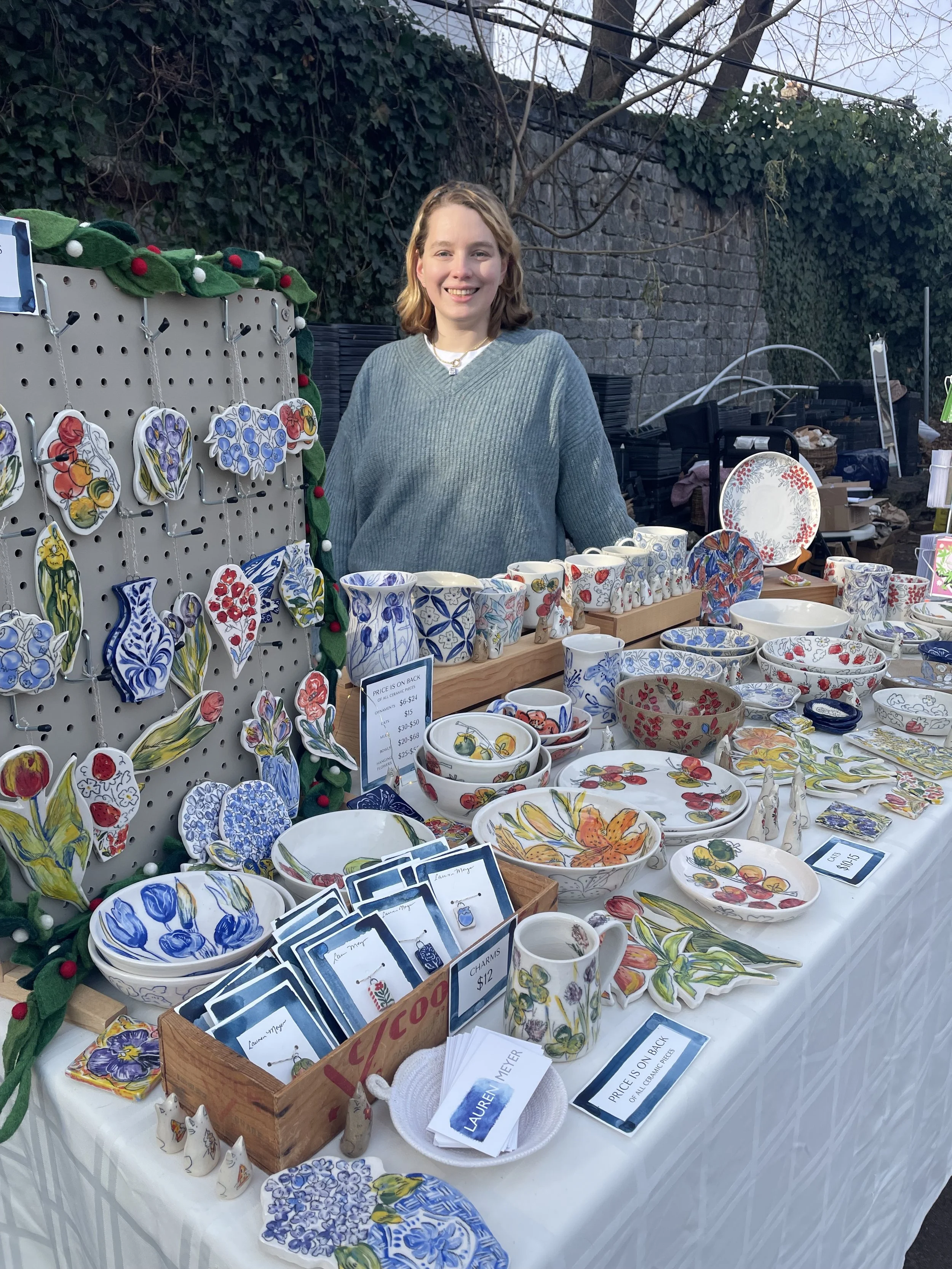 A woman smiling at a table with colorful ceramic dishware and decorations at an outdoor market stall.