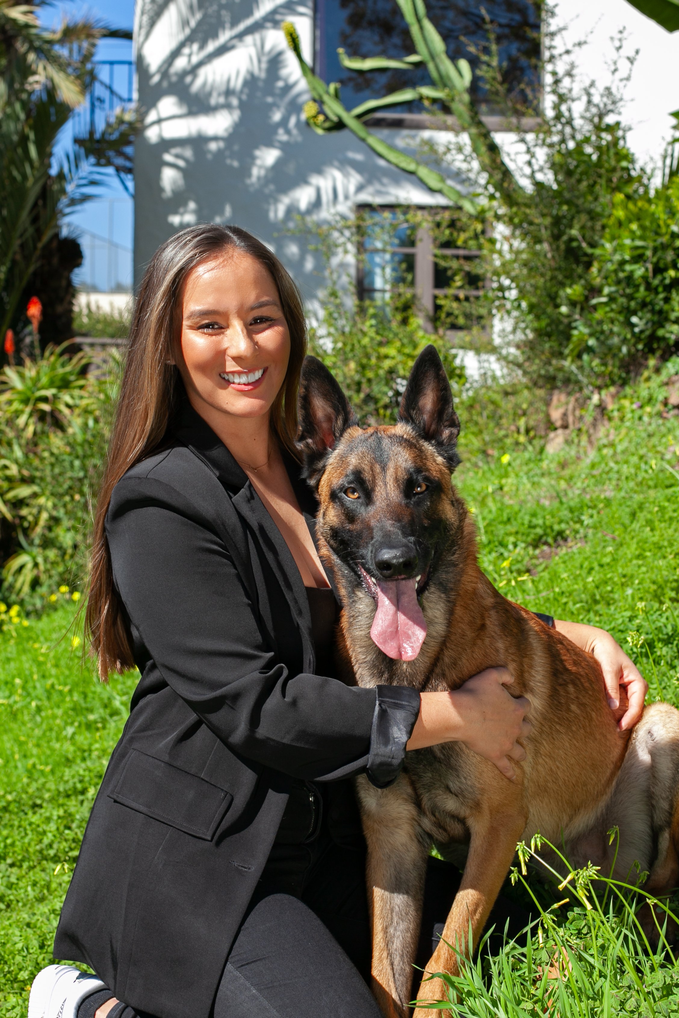A woman smiling and hugging a large, brown and black dog with pointed ears and tongue out outdoors on a sunny day.