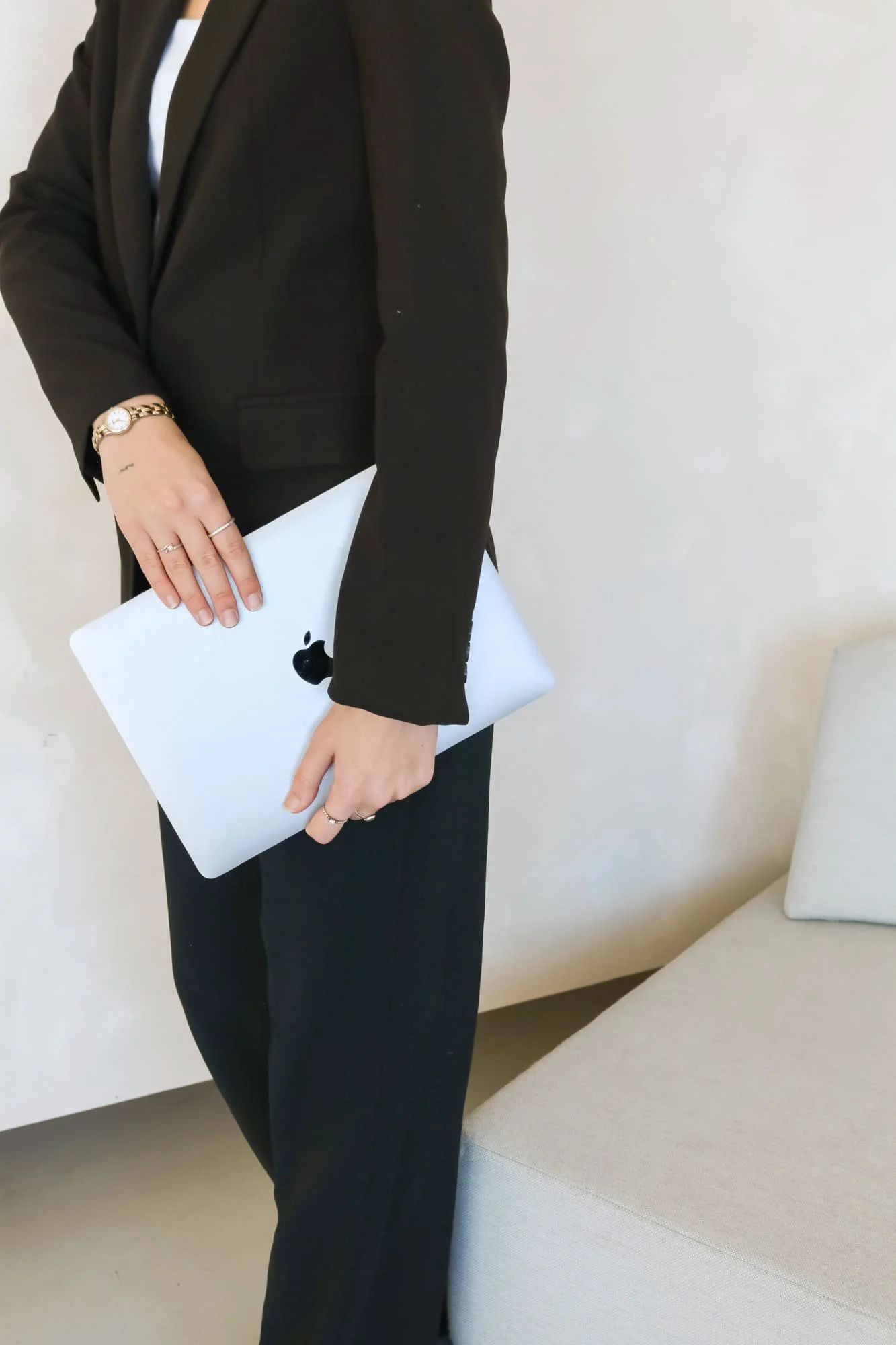 Une femme en complet noir tient un Macbook dans ces mains et posant dans un décor neutre.