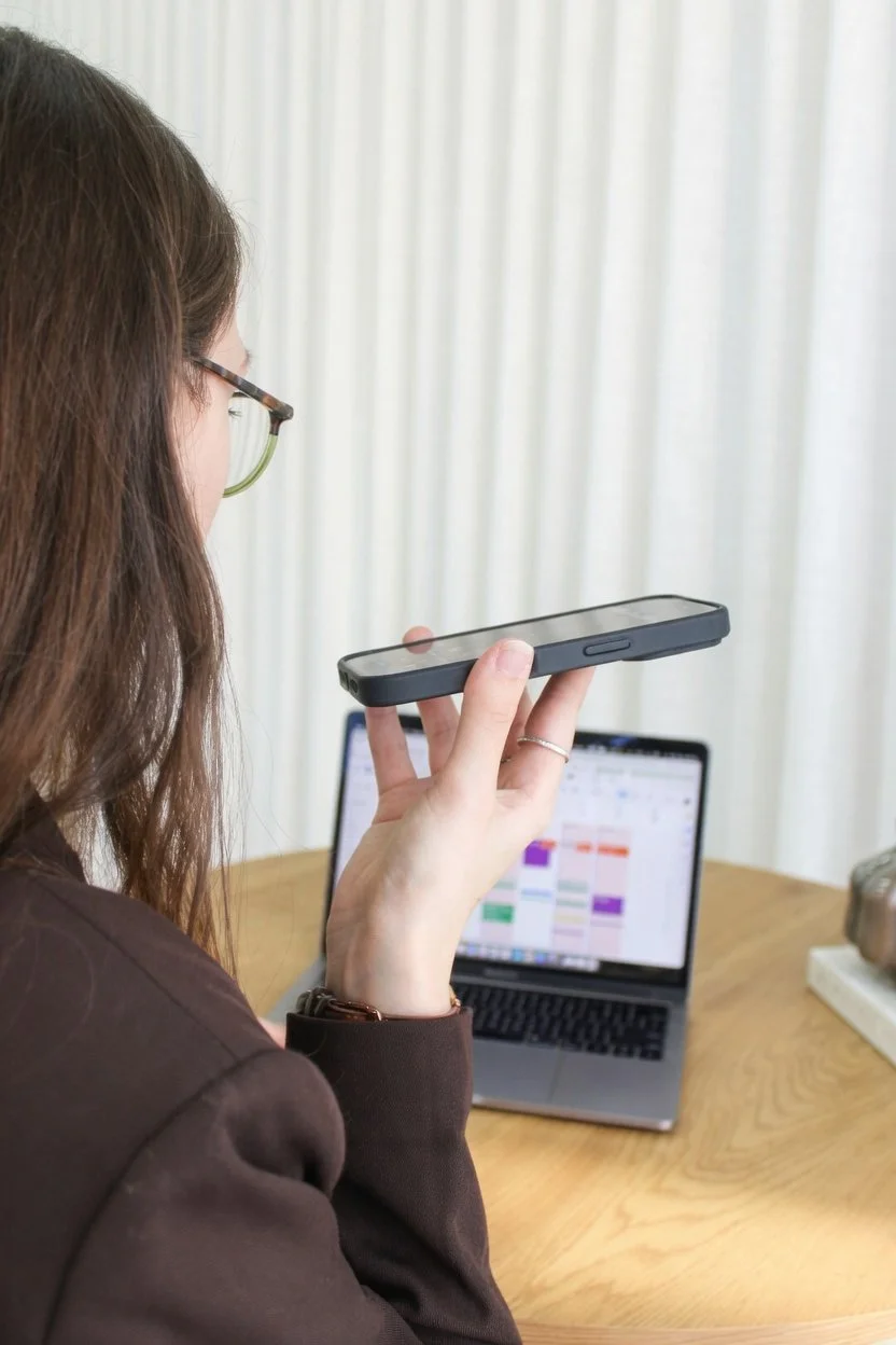 Femme à lunettes tenant un téléphone devant un MacBook affichant un calendrier coloré, sur une table en bois.