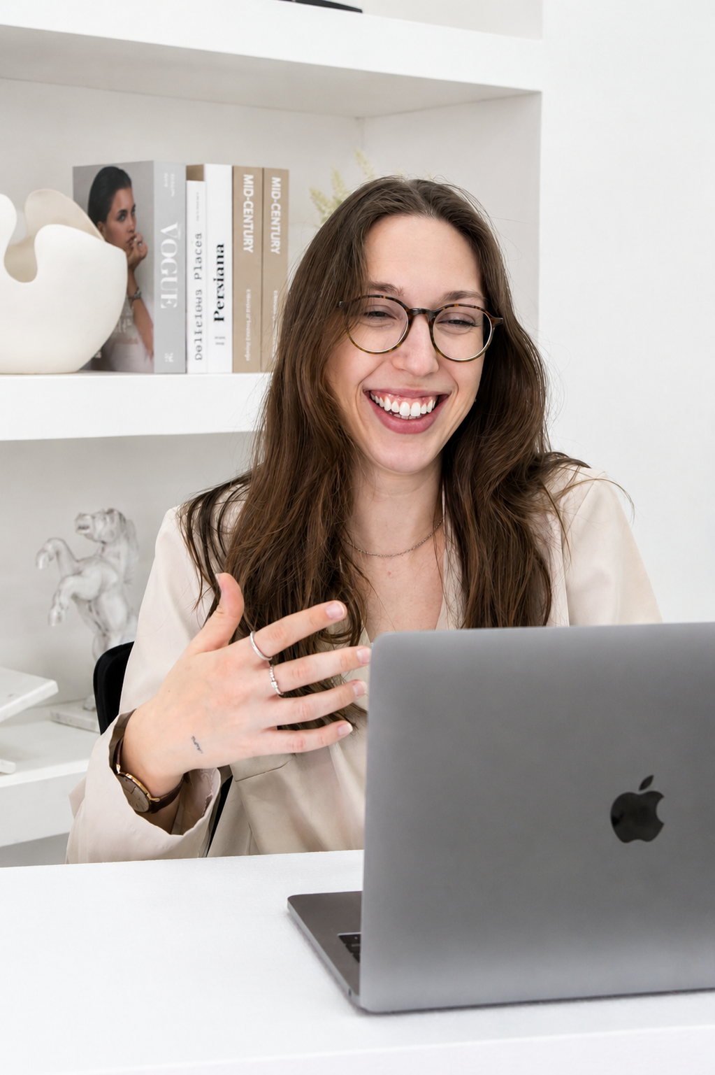 Abbie sourit en gesticulant devant un MacBook lors d'une visioconférence, dans un bureau blanc avec étagères et livres de décoration.
