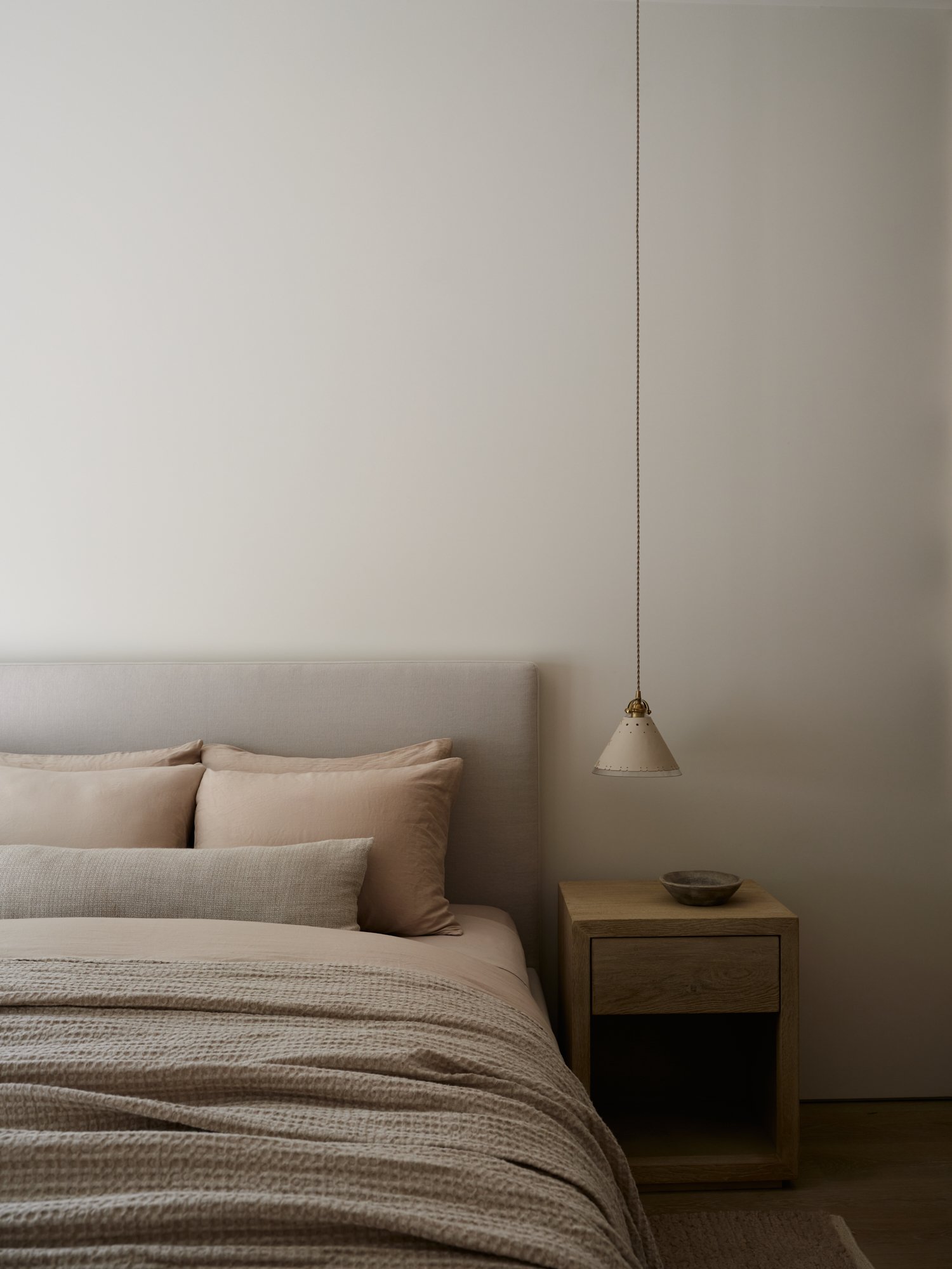Bedroom at The Stone House by Est. Collective, featuring beige Parachute bedding, layered textured linens, soft white plaster walls, a wooden nightstand with a shallow bowl, and a sculptural hanging pendant light.