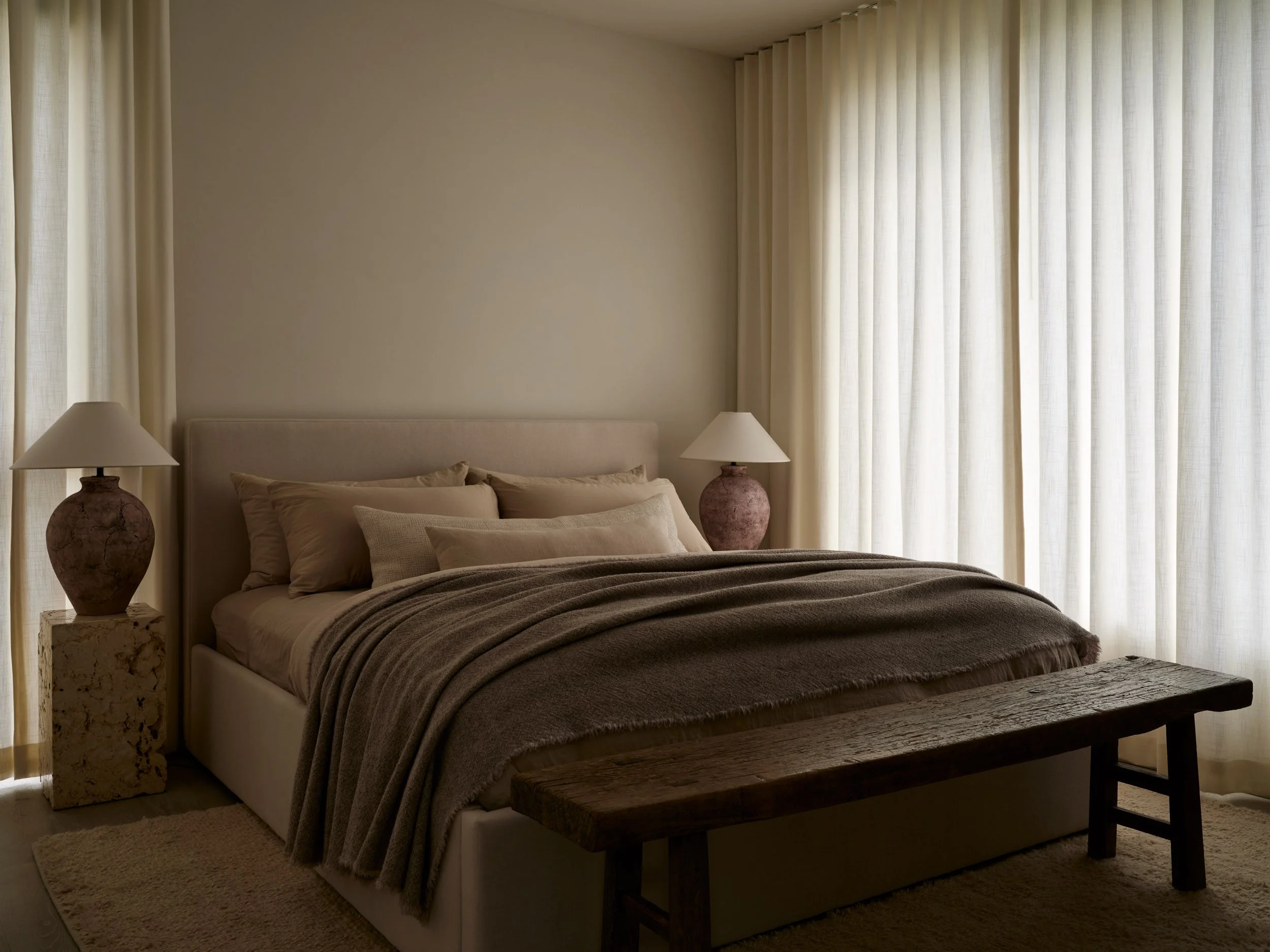 Bedroom at The Stone House by Est. Collective, featuring layered neutral linens, sculptural bedside lamps, a wooden bench, and soft natural textures.