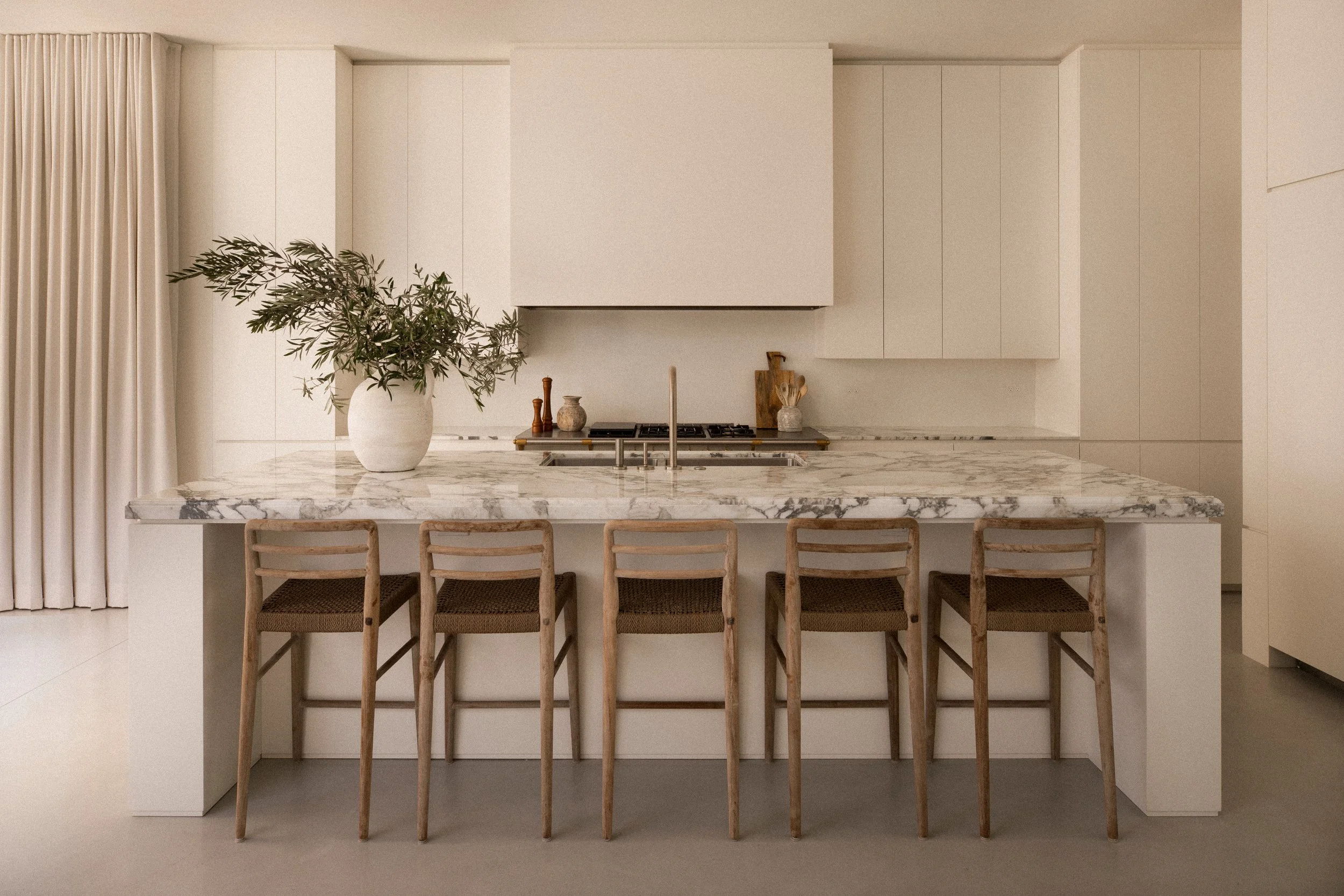 Modern kitchen with white cabinetry, marble countertop, and wooden bar stools. Decor includes a large potted plant, small vases, and a chopping board on the counter.
