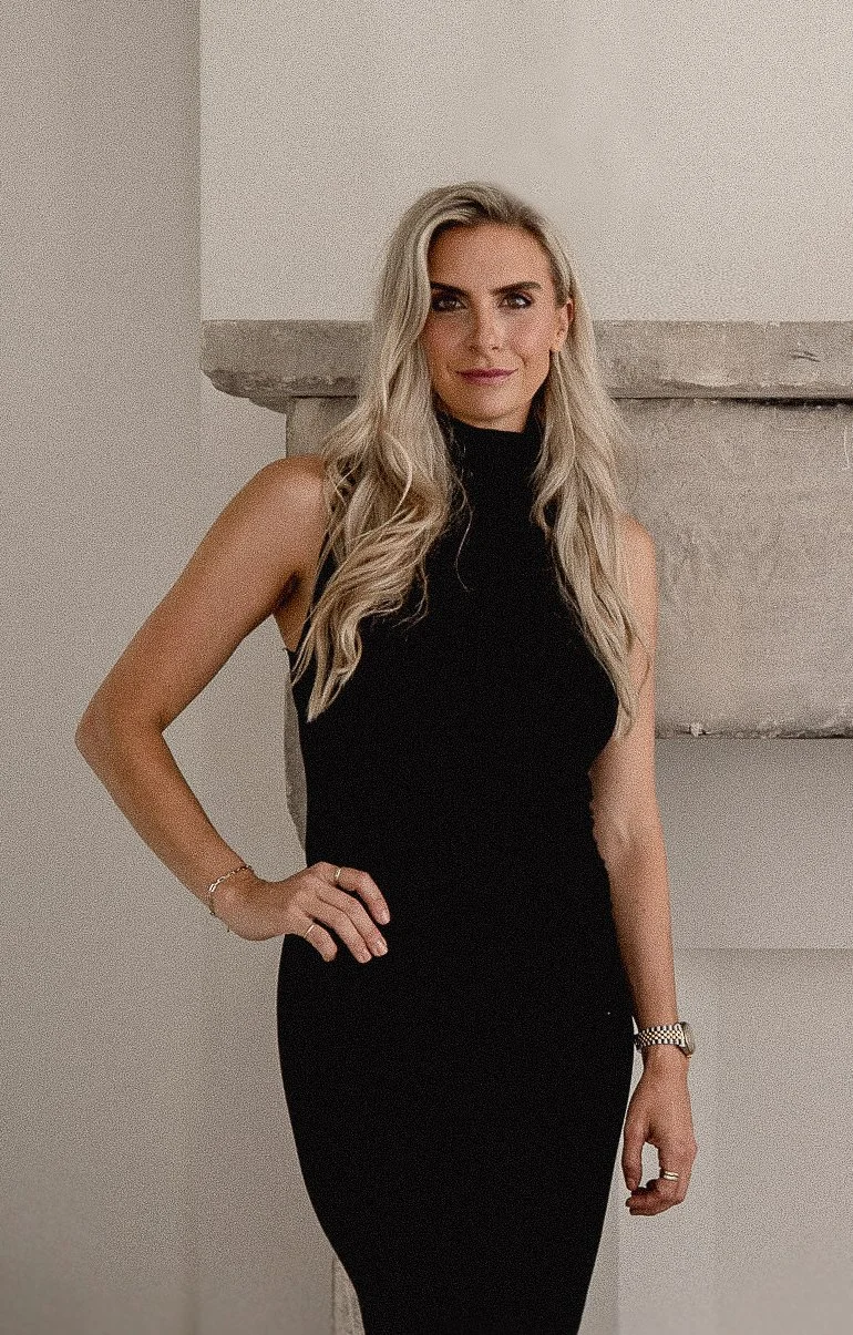 Nicole Heininger, founder of Est. Collective, standing in front of a neutral wall and fireplace wearing a black dress.