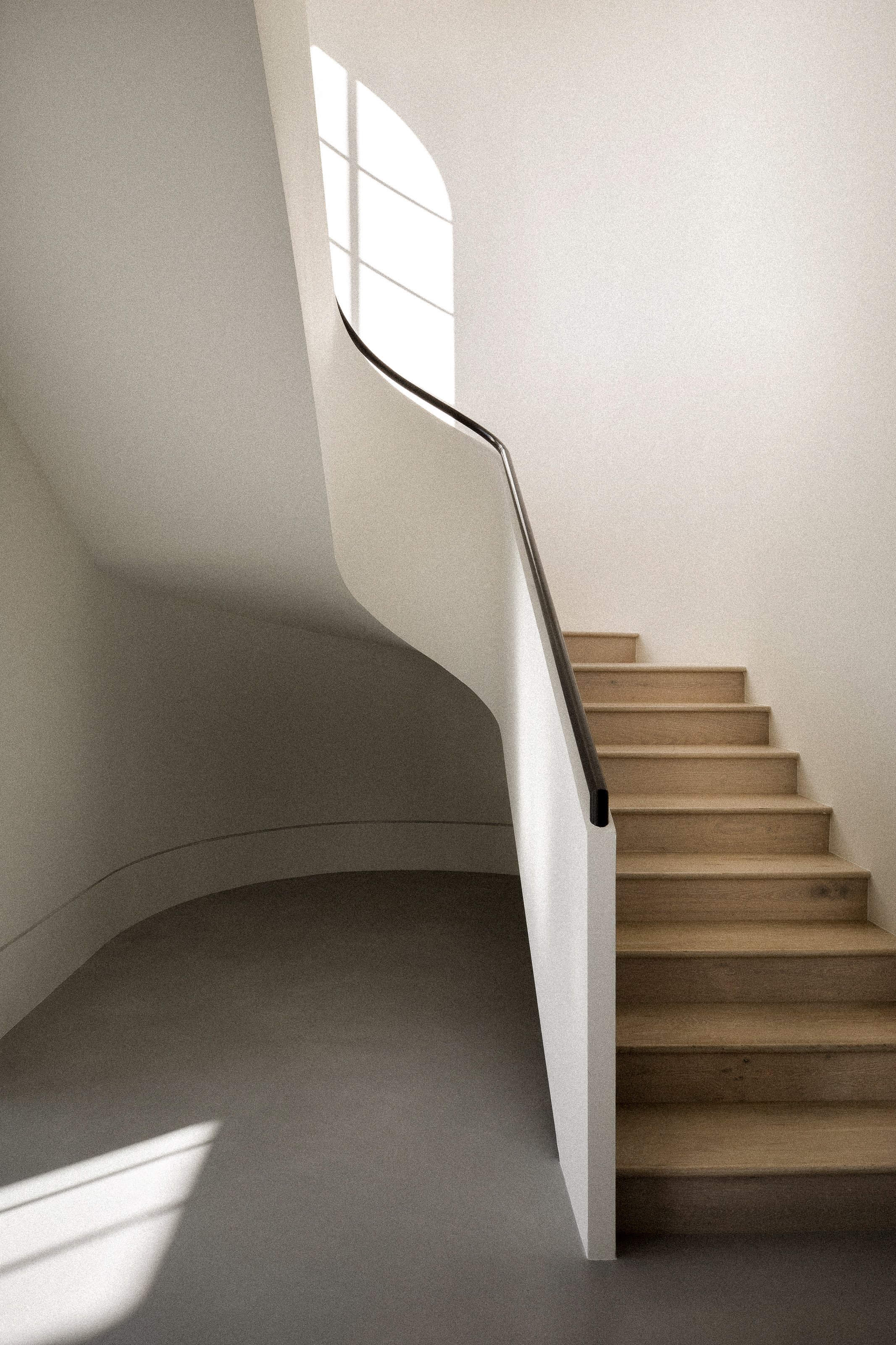 Interior view of a modern staircase with wooden steps, white walls, and a large window letting in natural light.