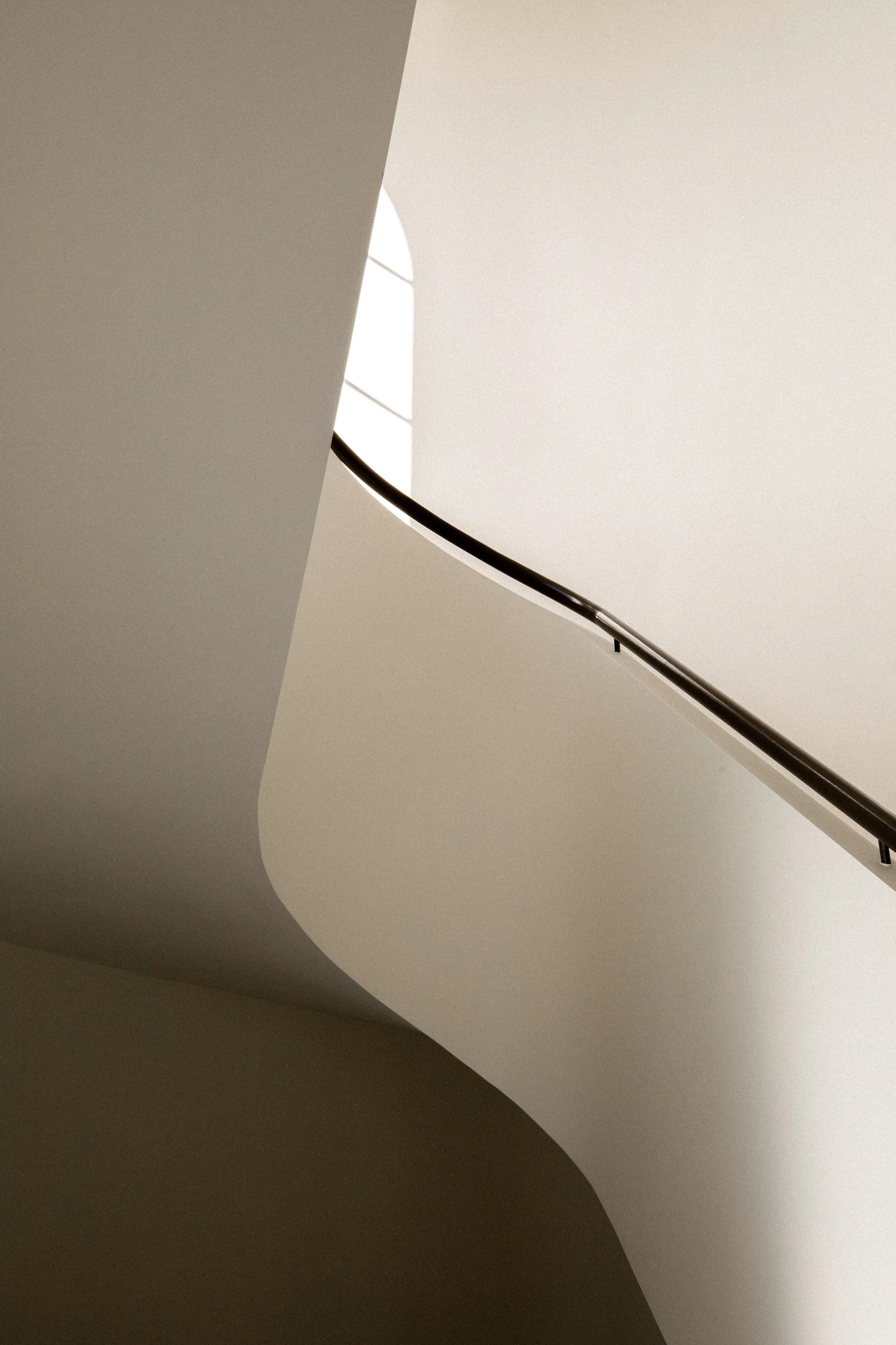 A minimalist staircase with white walls and a black handrail seen from below, with sunlight casting shadows.
