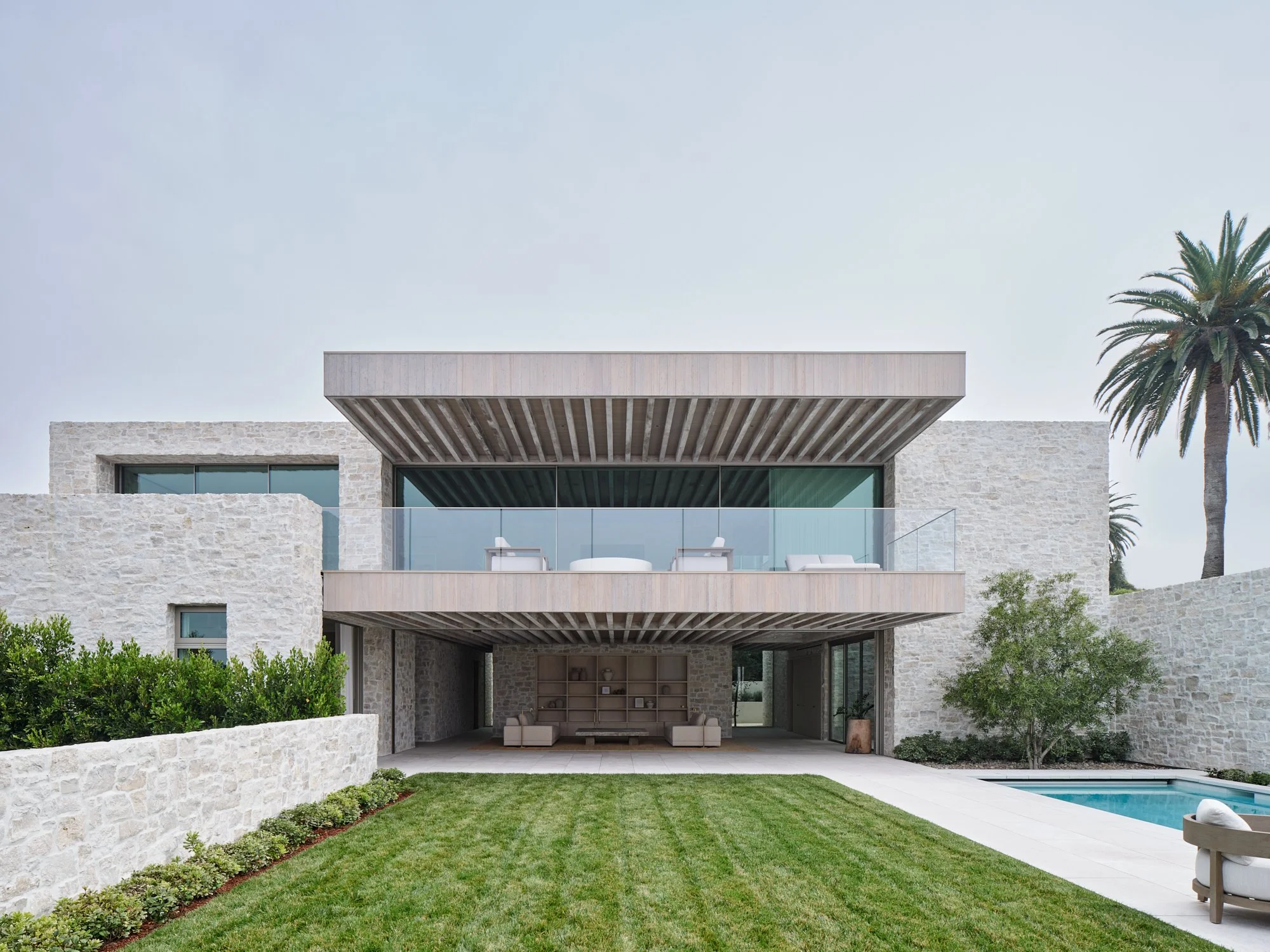 Modern house with stone exterior, large glass windows, two balconies, a backyard with a green lawn, a pool, and a view of a palm tree against a clear sky.