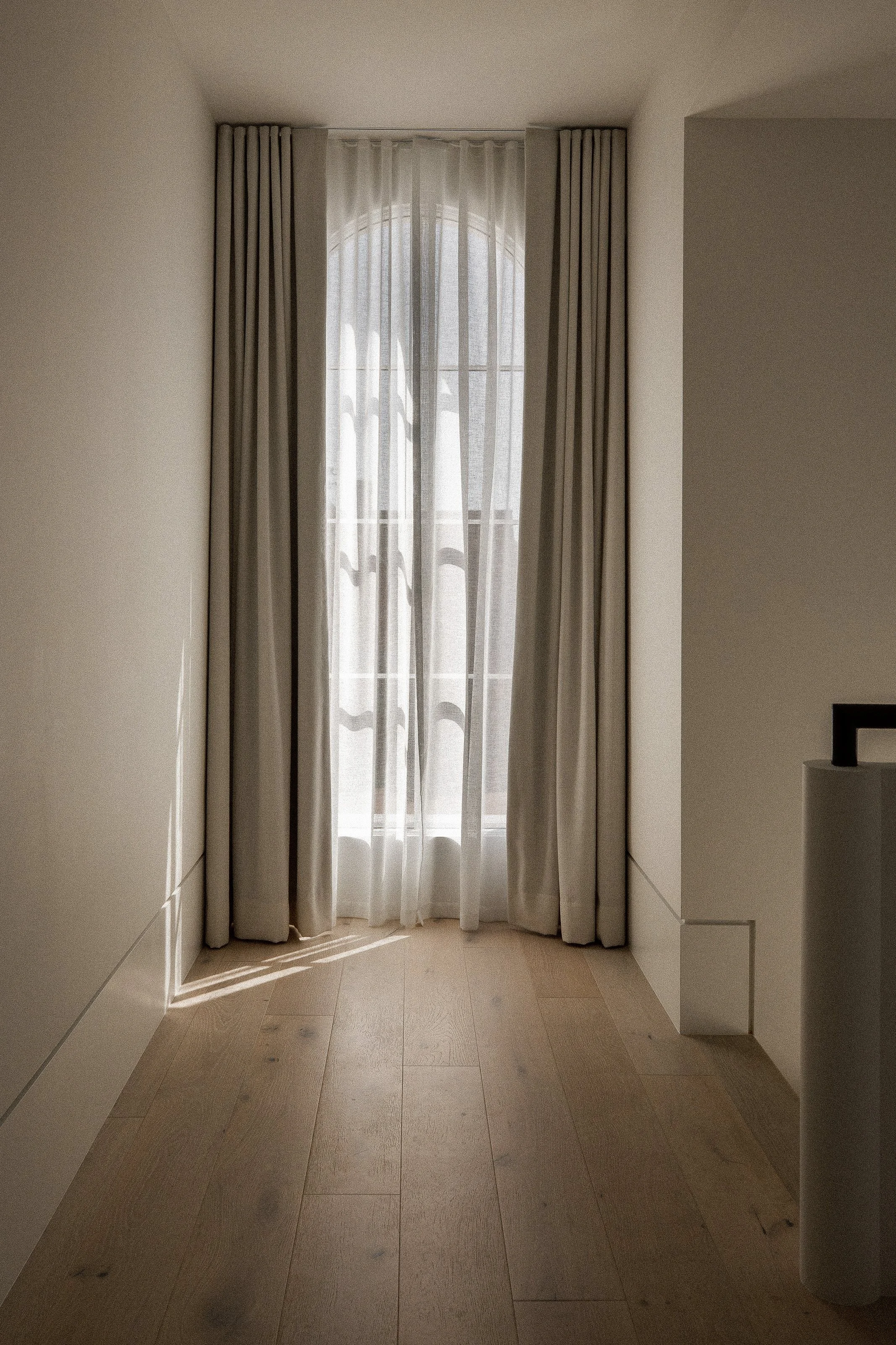 A window with layered sheer and opaque curtains in a bright, neutral-colored room, with sunlight casting shadows on the hardwood floor.