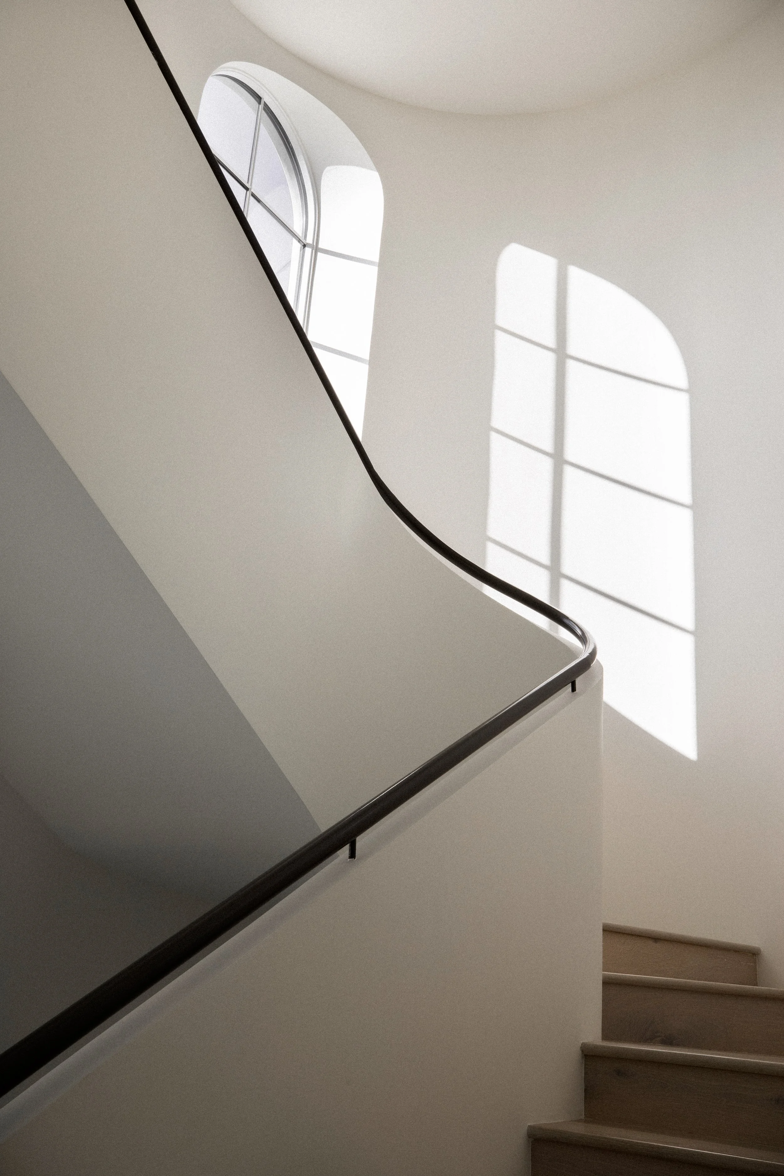 Interior view of a staircase with wooden steps, black railing, and sunlight streaming through arched windows, casting shadows on the wall.