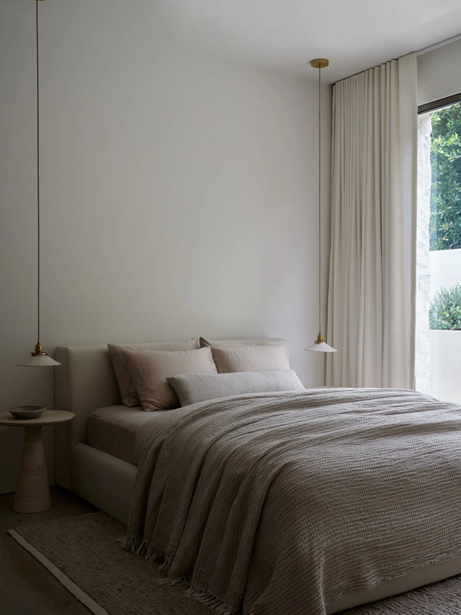 A neatly made bed with beige linens and pillows, two hanging pendant lights on either side, a side table with a bowl, and a large window with white curtains overlooking greenery.