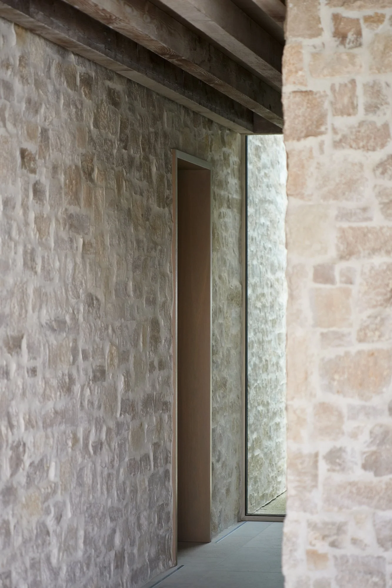 A modern interior corridor with stone walls, a wooden ceiling, and a large mirror on the right side, with a door visible at the end.