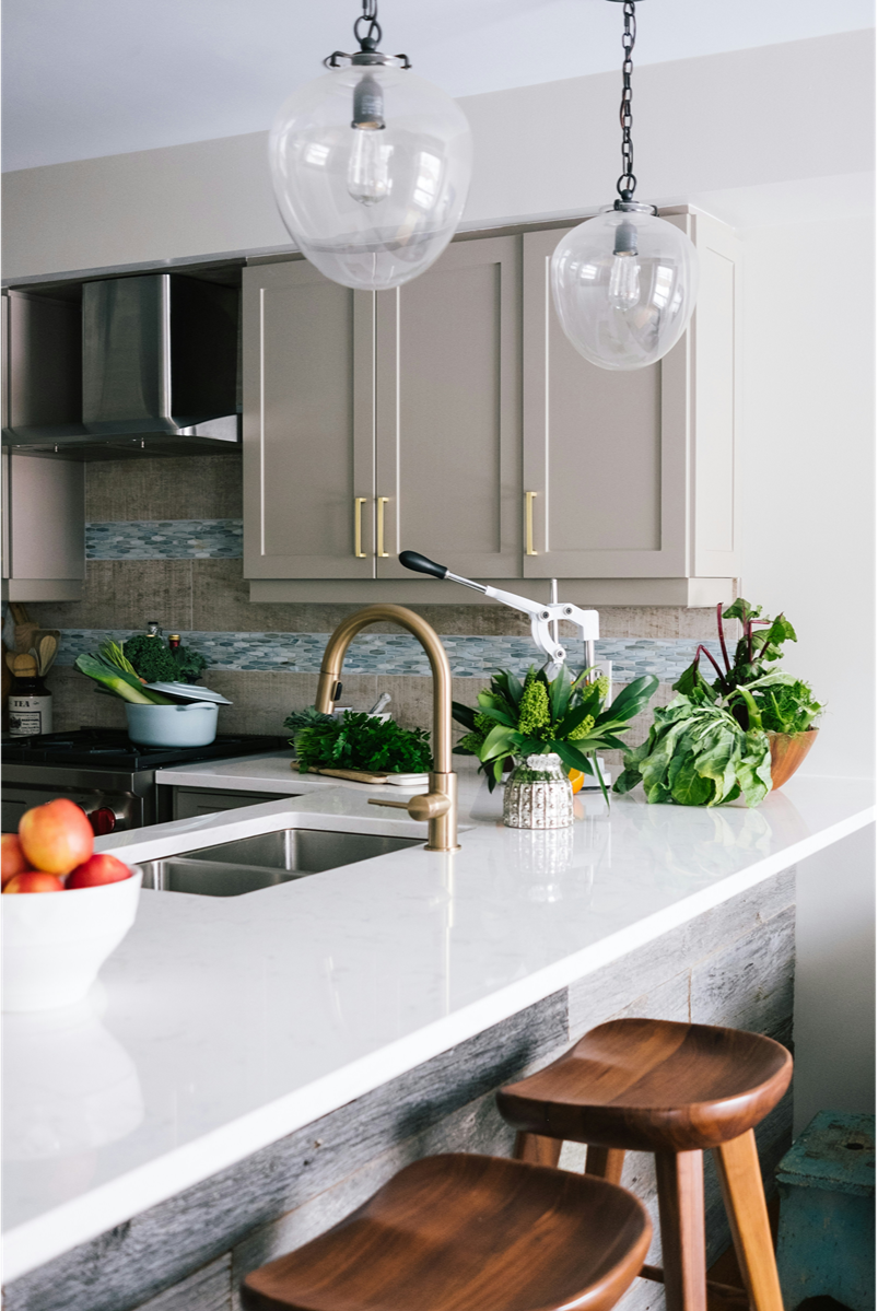 Modern kitchen with beige cabinets, white countertop, stainless steel appliances, and hanging glass pendant lights. There are green plants and a bowl of apples on the counter, with two wooden stools at the breakfast bar.