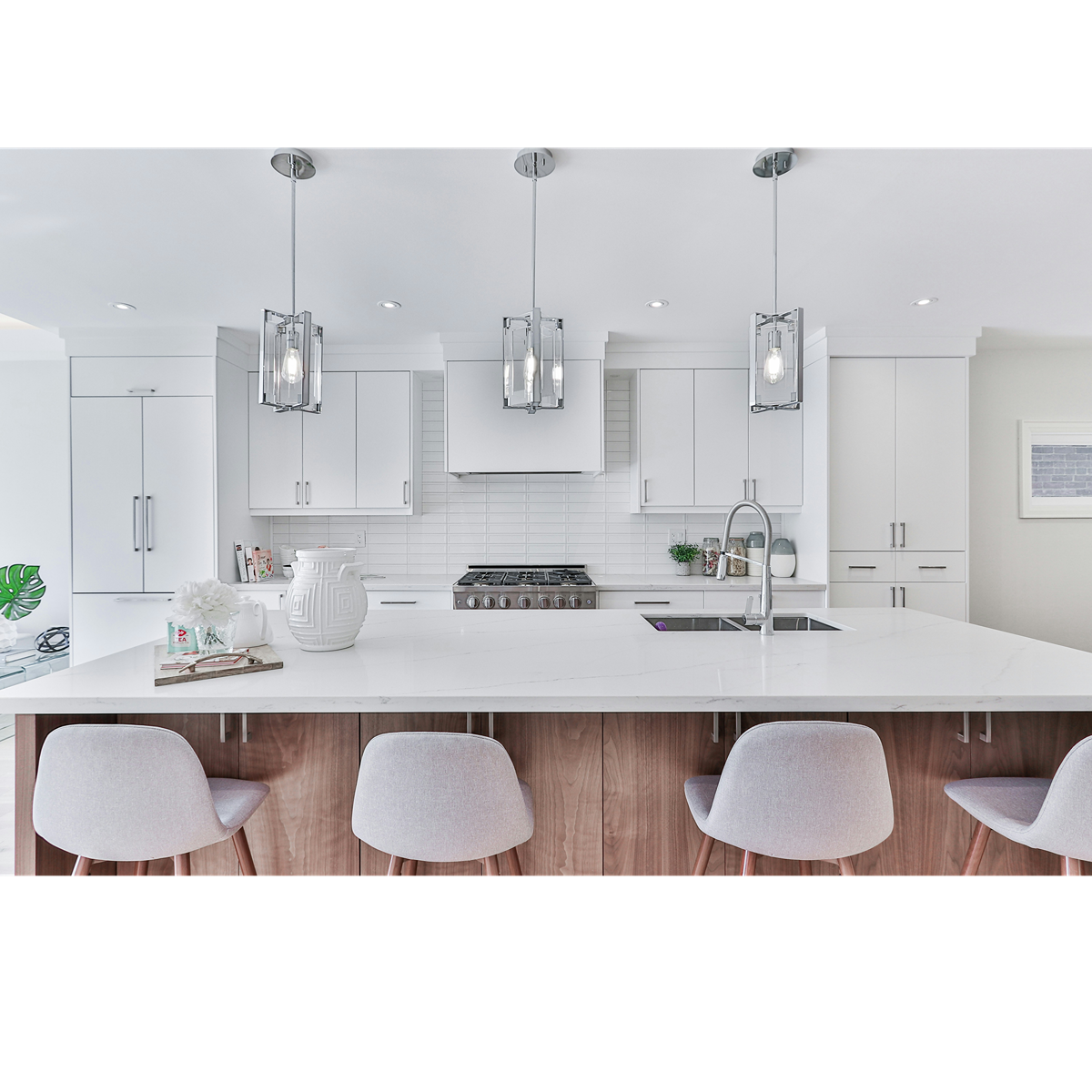 Modern white kitchen with a large breakfast bar, three grey chairs, pendant lights, white cabinets, a stove, and a kitchen sink.