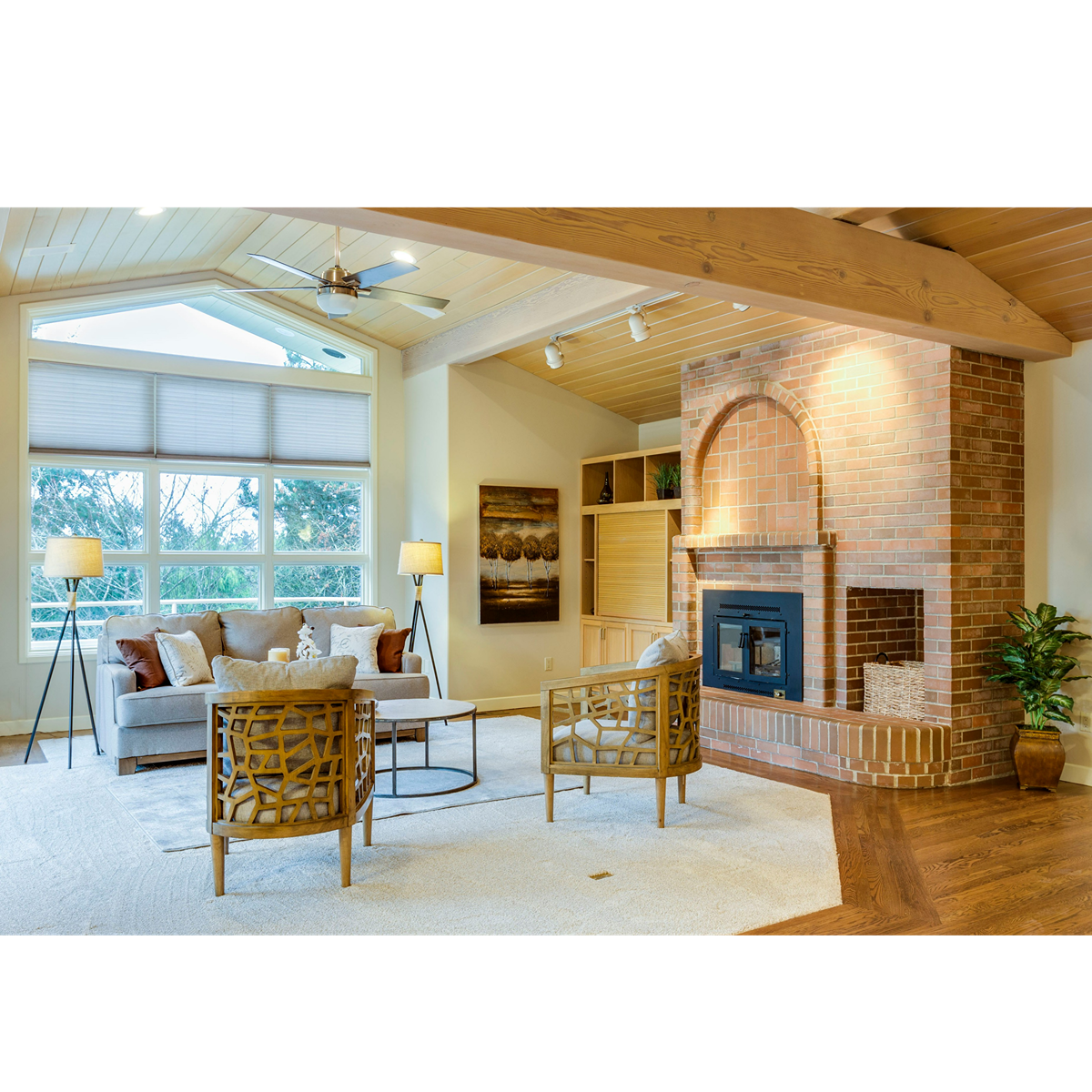 Living room with large windows, beige sofa with pillows, two floor lamps, wooden chairs, brick fireplace, wooden ceiling, ceiling fan, artwork, and plants.