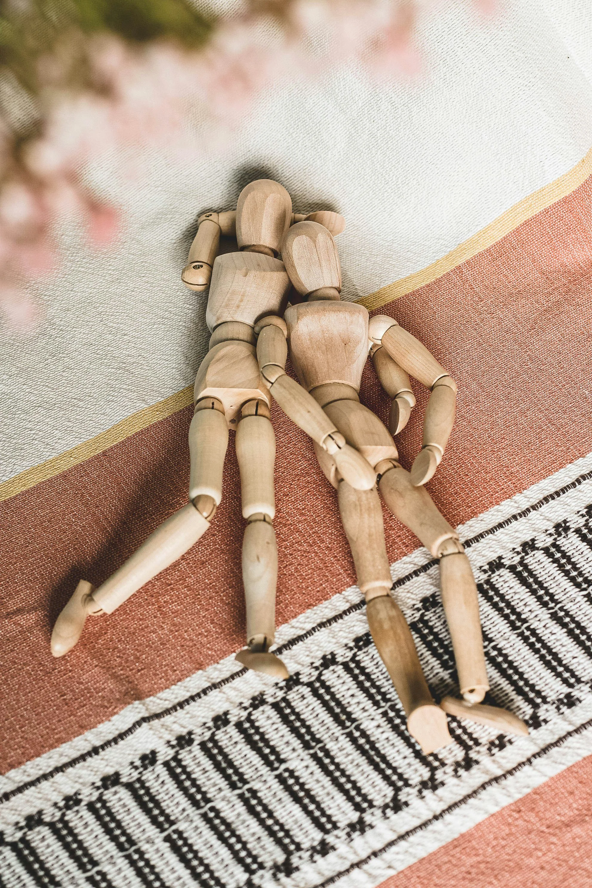 A wooden mannequin lying on a patterned textile surface, partially visible with pink flowers blurred in the foreground.