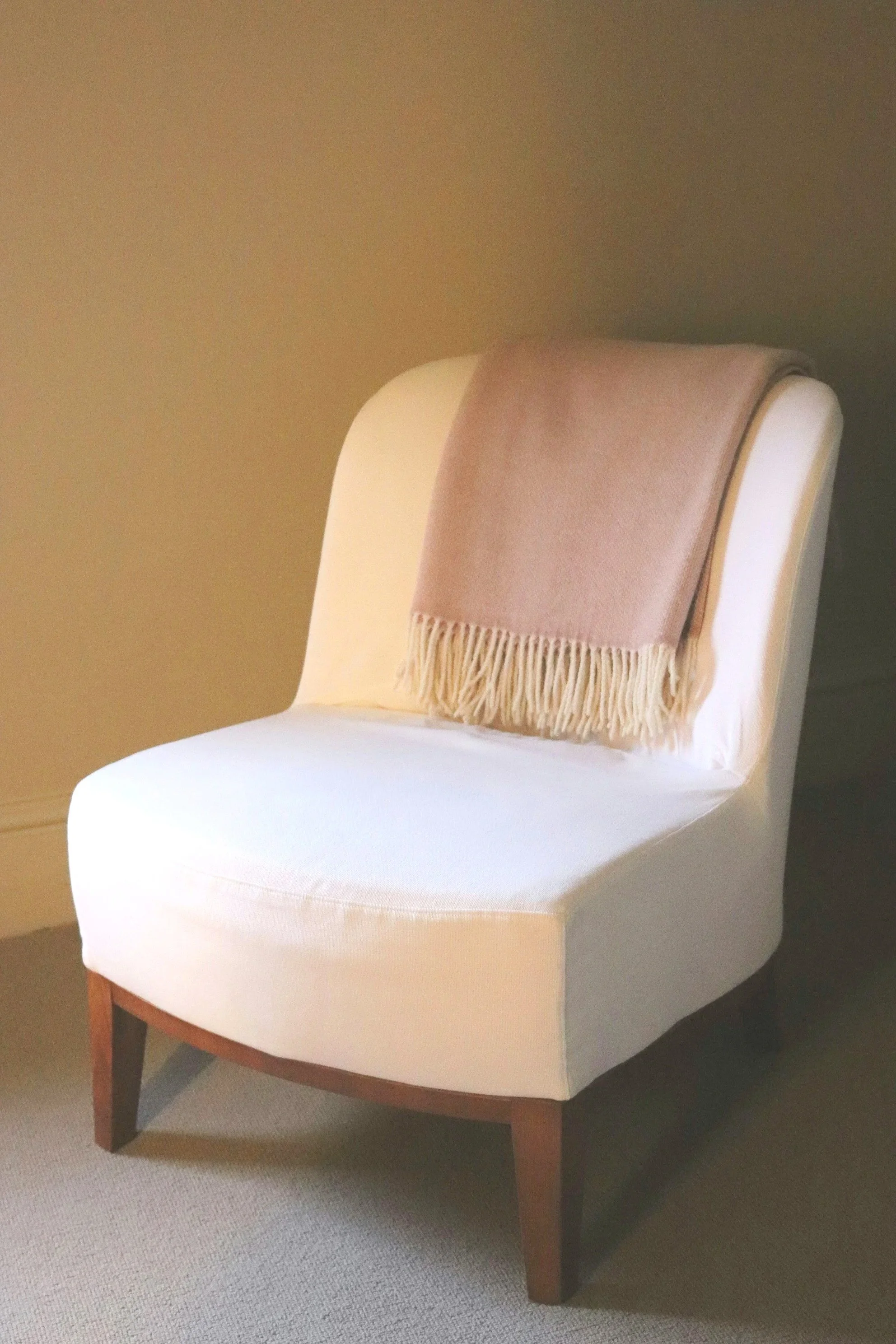 White dining chair with wooden legs and a beige fringe scarf draped over the backrest, placed on a carpeted floor against a plain wall.