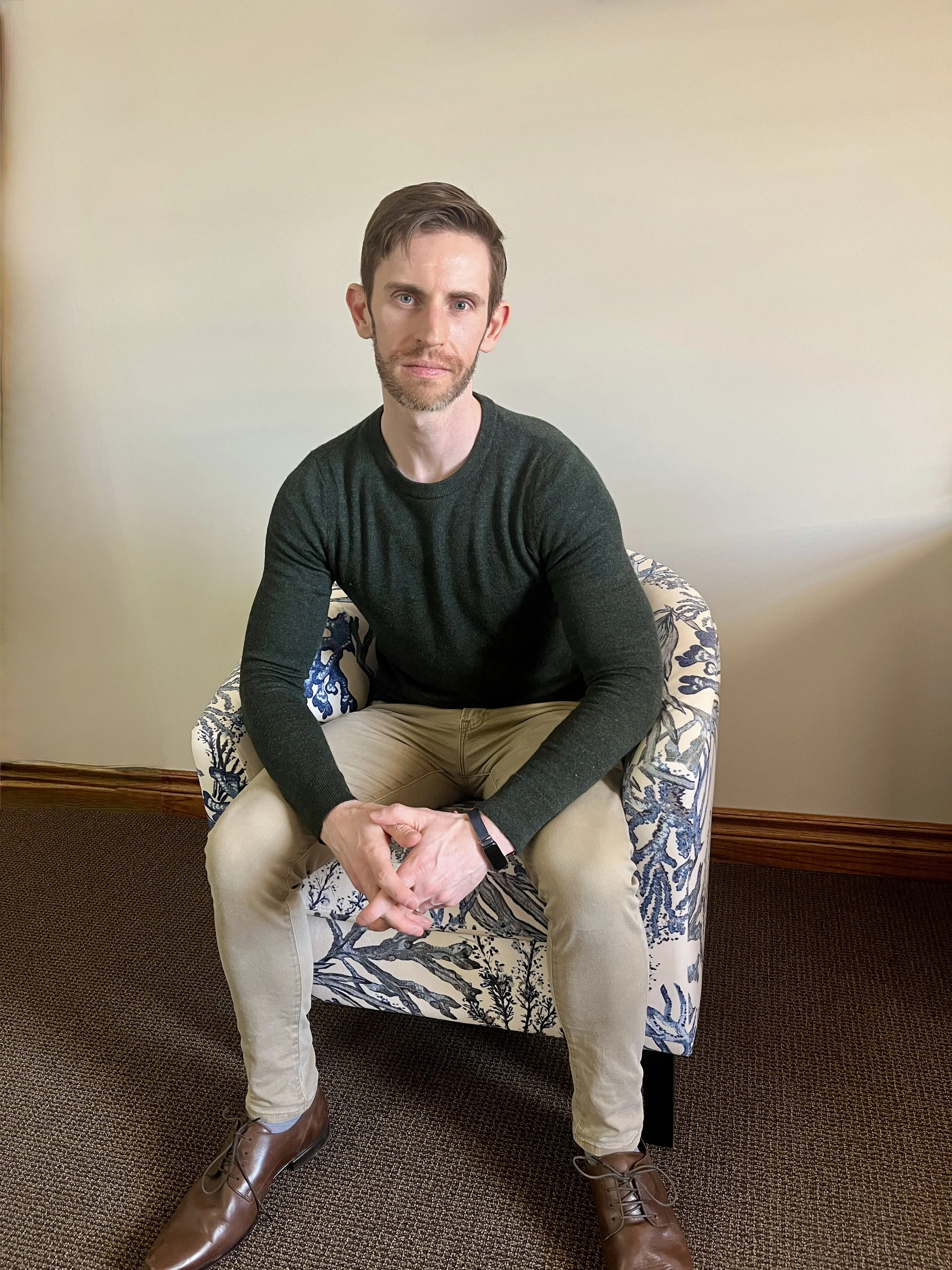 A man with short dark hair and a beard sitting on a patterned armchair in a room with beige walls and a brown carpet. He is wearing a long-sleeved dark green shirt, cream-colored pants, and brown shoes, with his hands clasped on his lap.