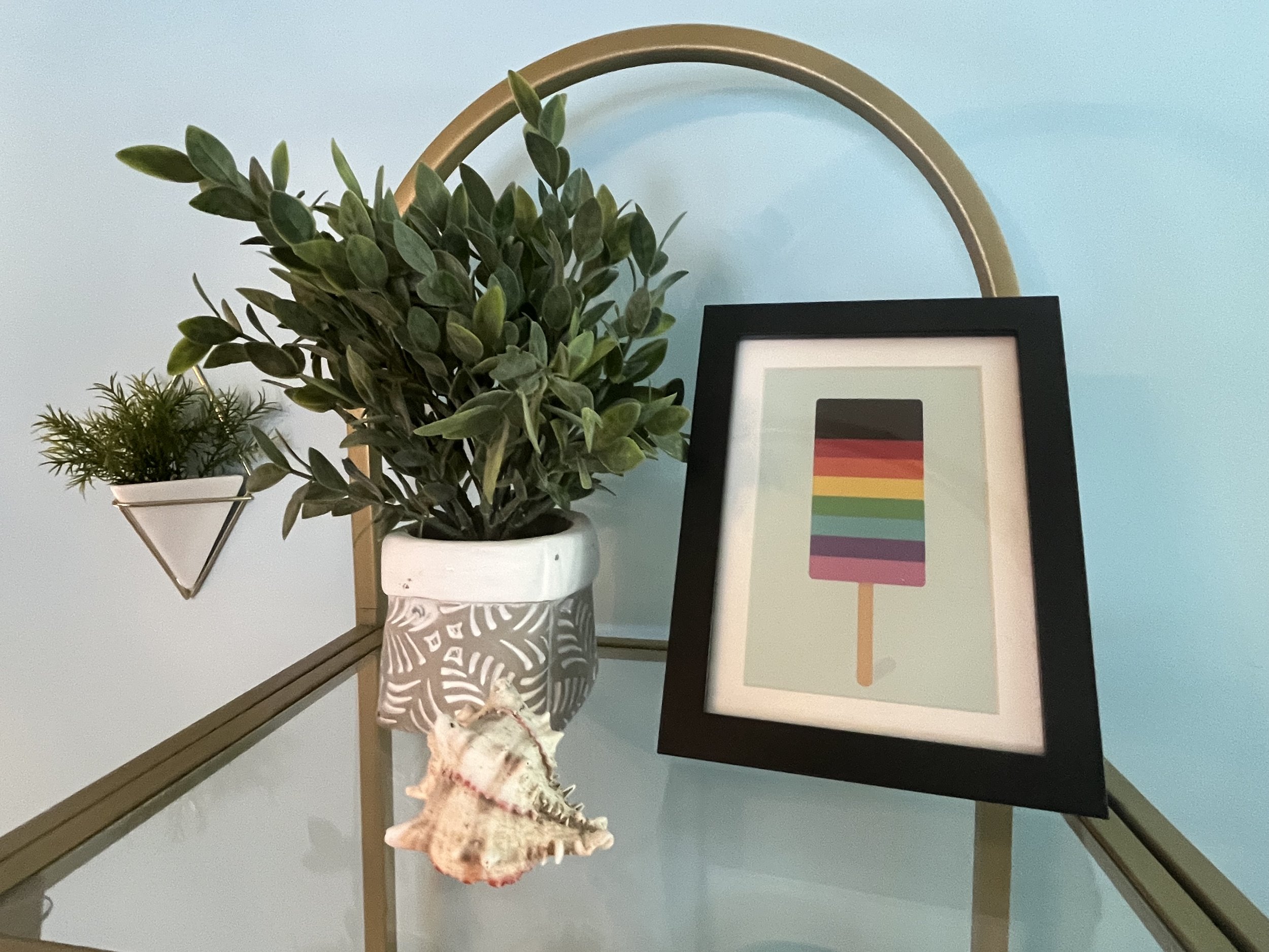 Decorative surface with potted plants, a framed picture of a popsicle, and a seashell, all arranged on a table against a light blue wall.