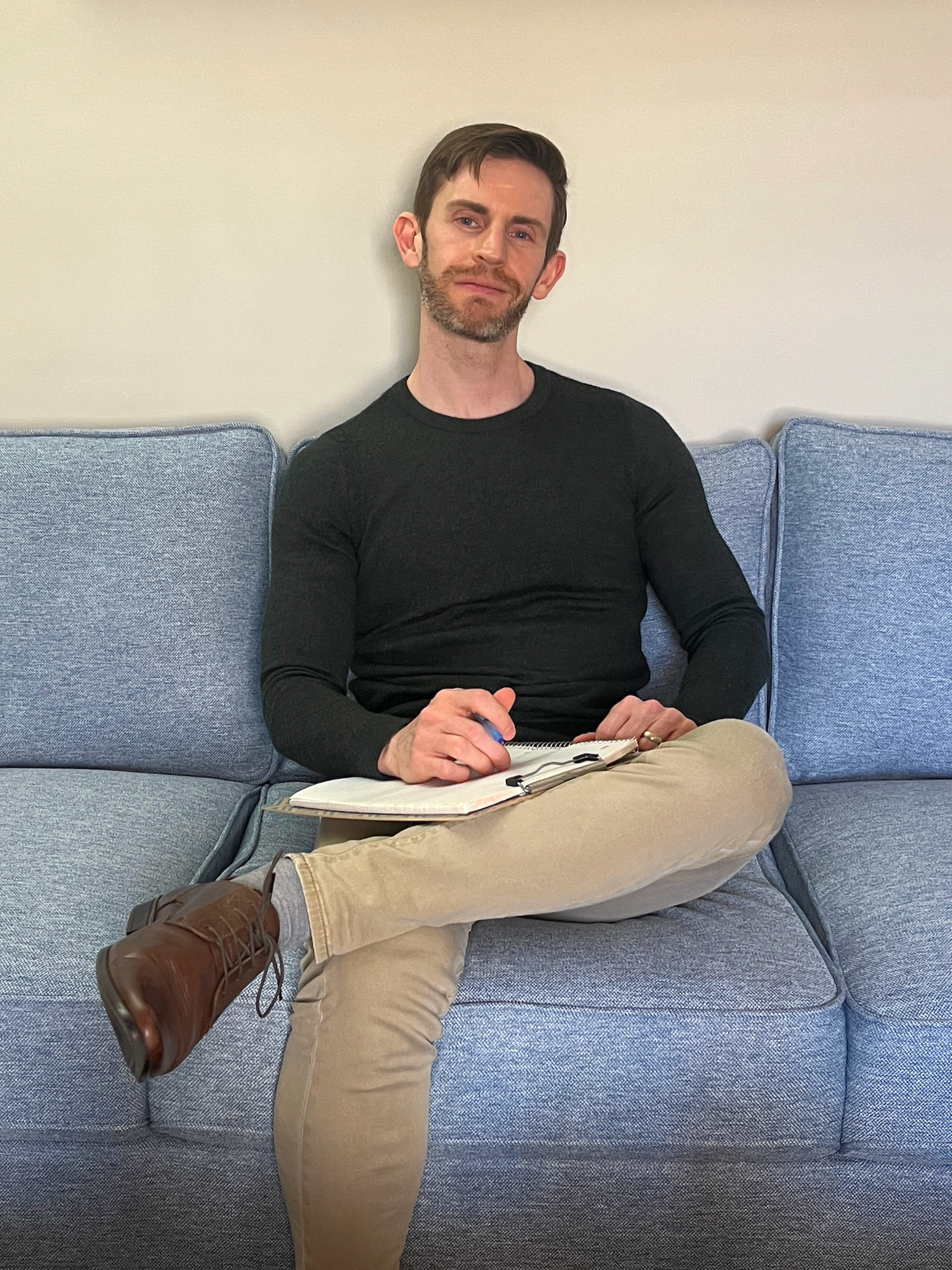 A man with brown hair, beard, and mustache, sitting on a blue sofa, taking notes in a notebook with a pen, wearing a black long-sleeve shirt, khaki pants, and brown shoes, against a plain light-colored wall.