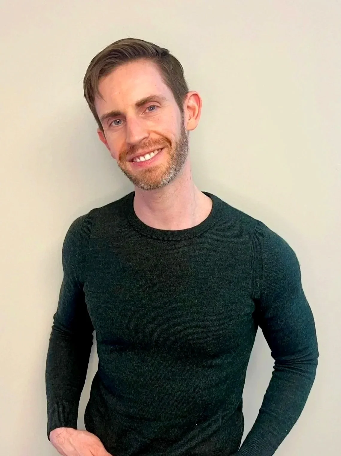 A man with short brown hair, beard, and blue eyes smiling, wearing a dark, long-sleeved shirt, standing against a plain, light-colored wall.