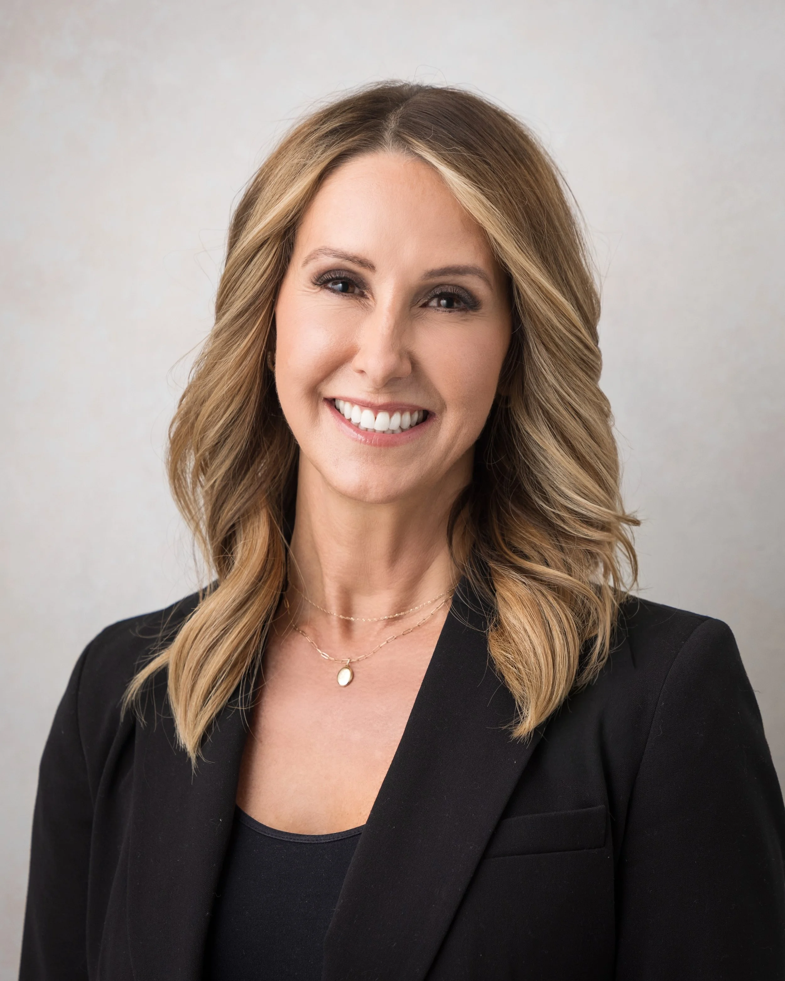 Professional woman with shoulder-length blonde hair, smiling, wearing a black blazer and a delicate gold necklace, standing against a neutral background.