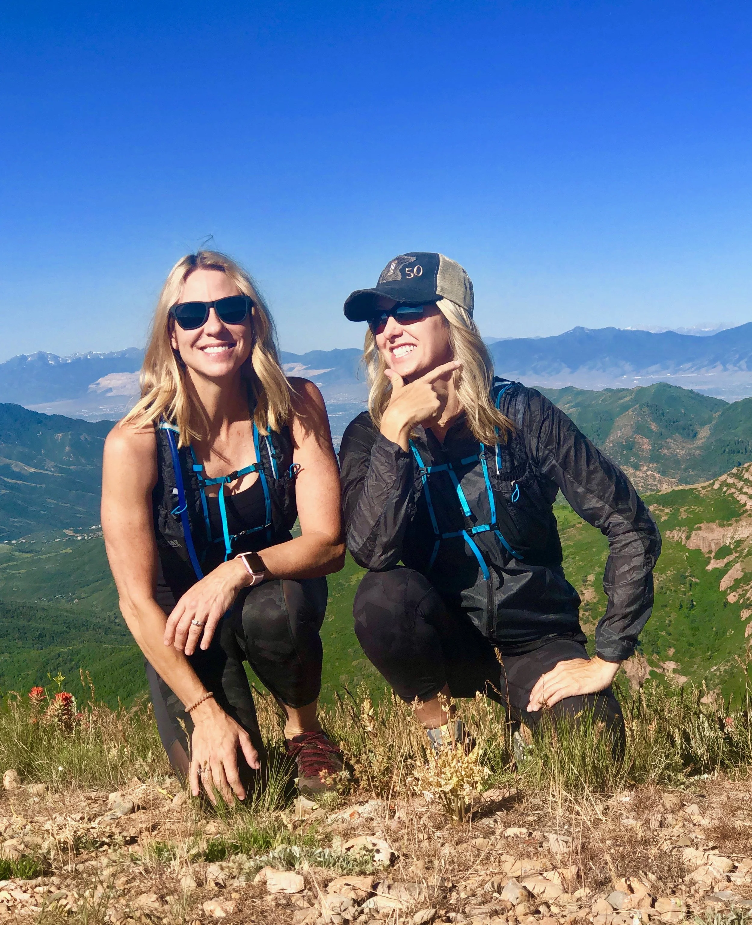 Two women on a mountain hike, smiling and striking playful poses with a scenic mountain landscape in the background.
