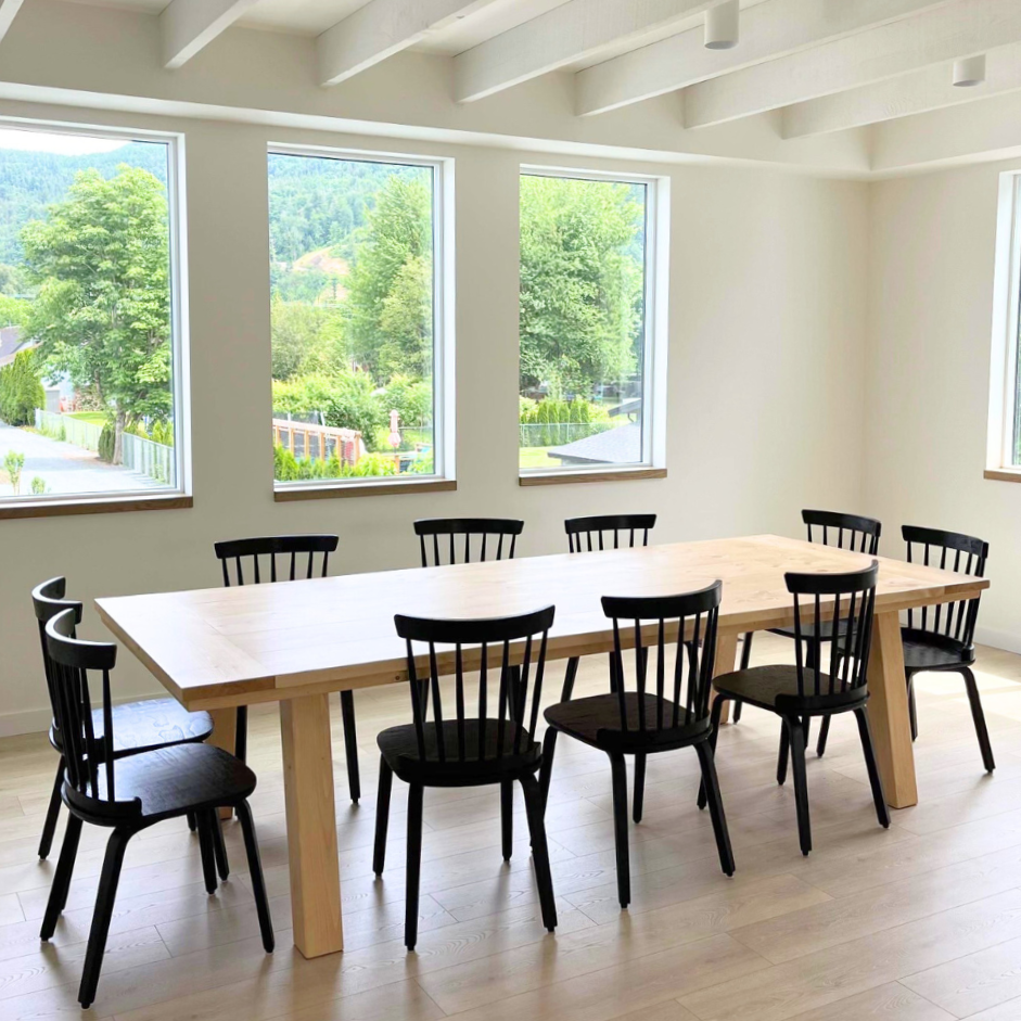 A Scandinavian dining table made of wood with sleek, off-centered legs, surrounded by black chairs in a bright dining room setting.