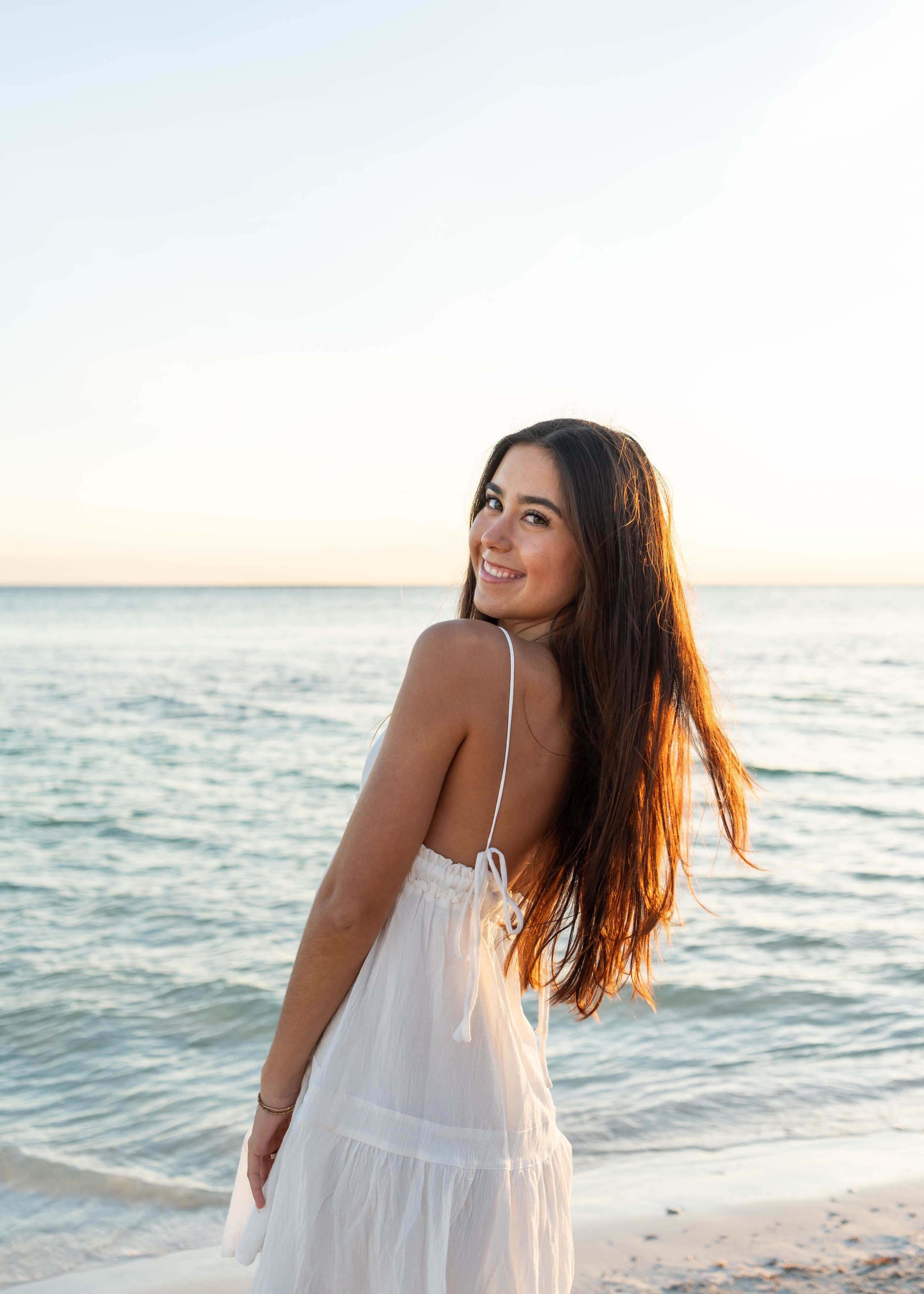 Smiling woman with long brown hair in a white dress at the beach during sunset.