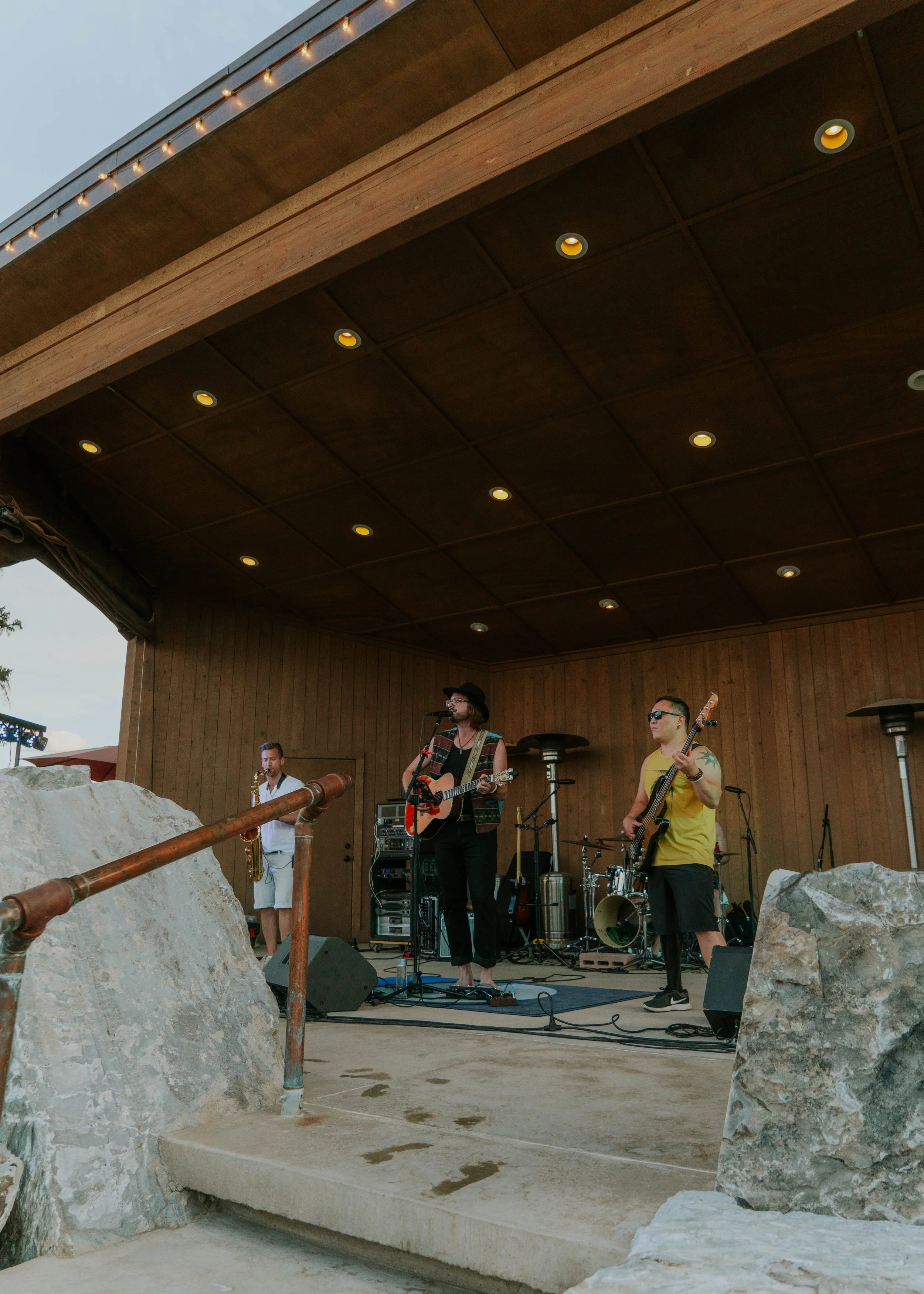 A band performing on an outdoor wooden stage, with three musicians visible: a singer and guitarist, a bass player, and a saxophonist, with stage equipment and large rocks in foreground.