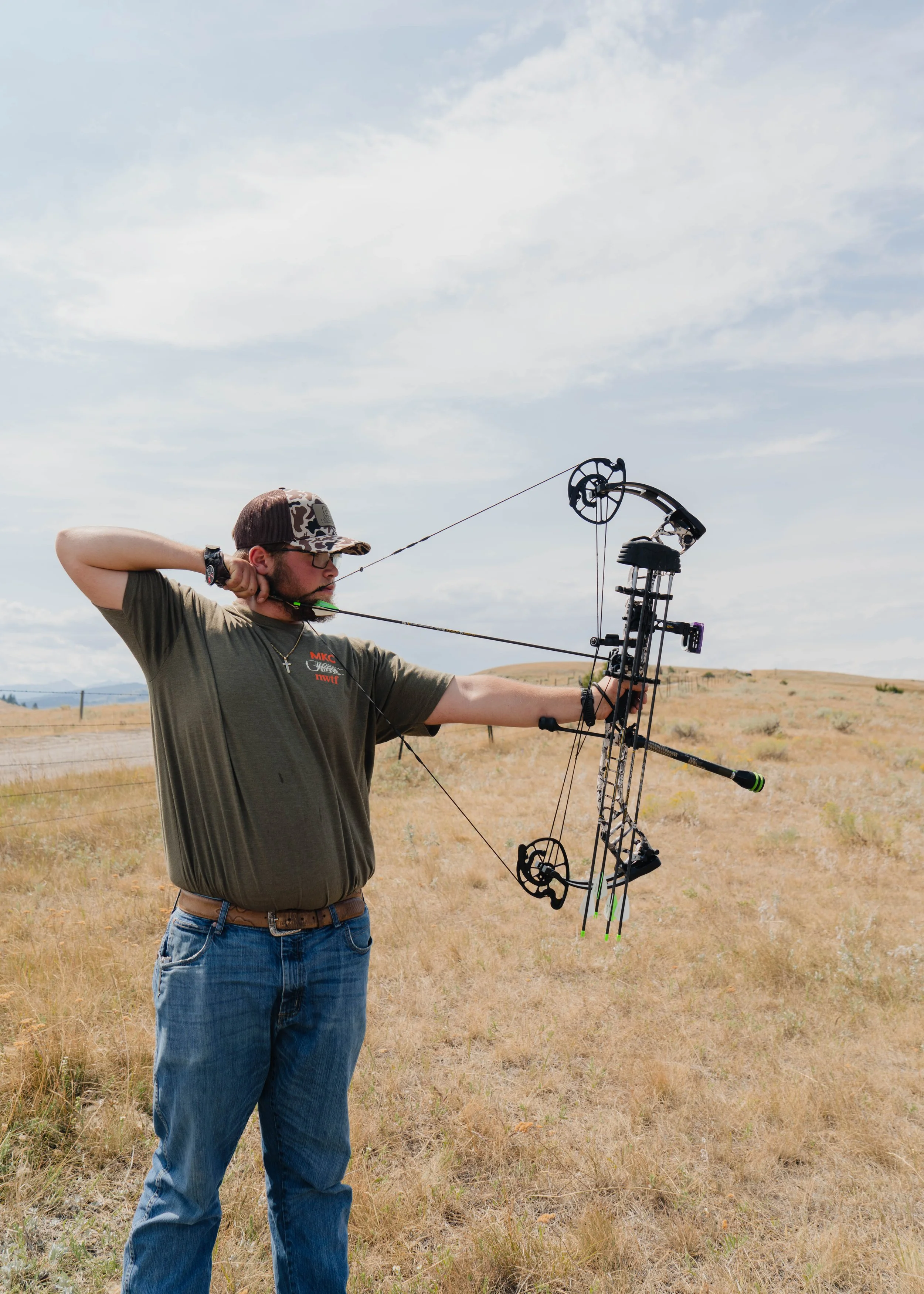 A man practicing archery outdoors on a sunny day in a grassy field, aiming with a modern compound bow.