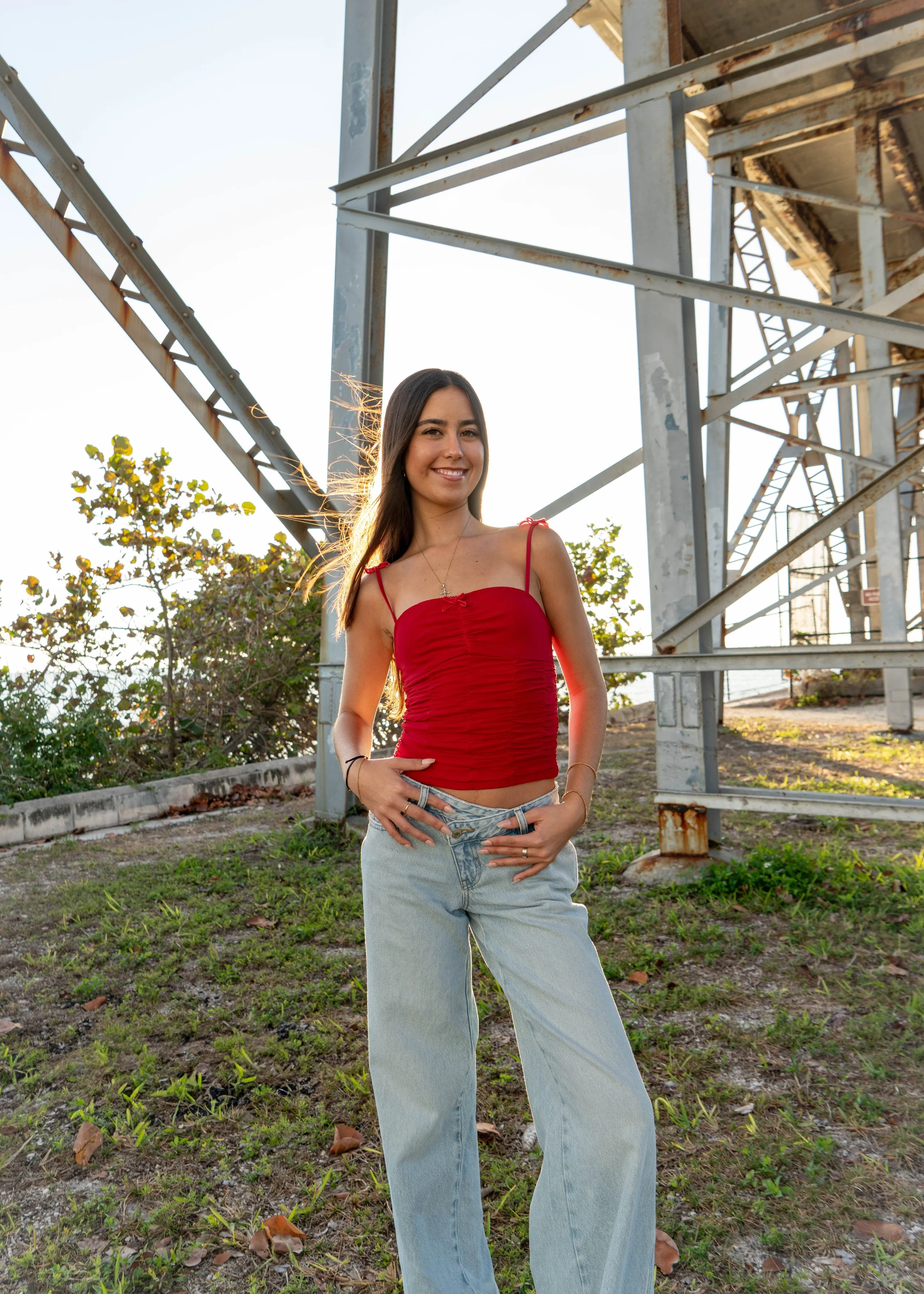 Young woman standing outdoors near a steel structure, wearing a red spaghetti strap top and light-wash jeans, smiling at the camera with trees and blue sky in the background during sunset.