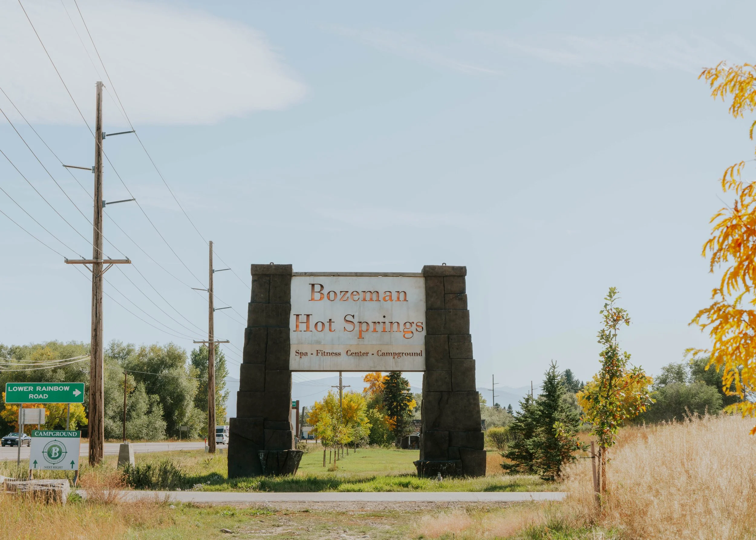 Entrance sign for Bozeman Hot Springs, featuring details for spa, fitness center, and campground, with trees and blue sky in the background.