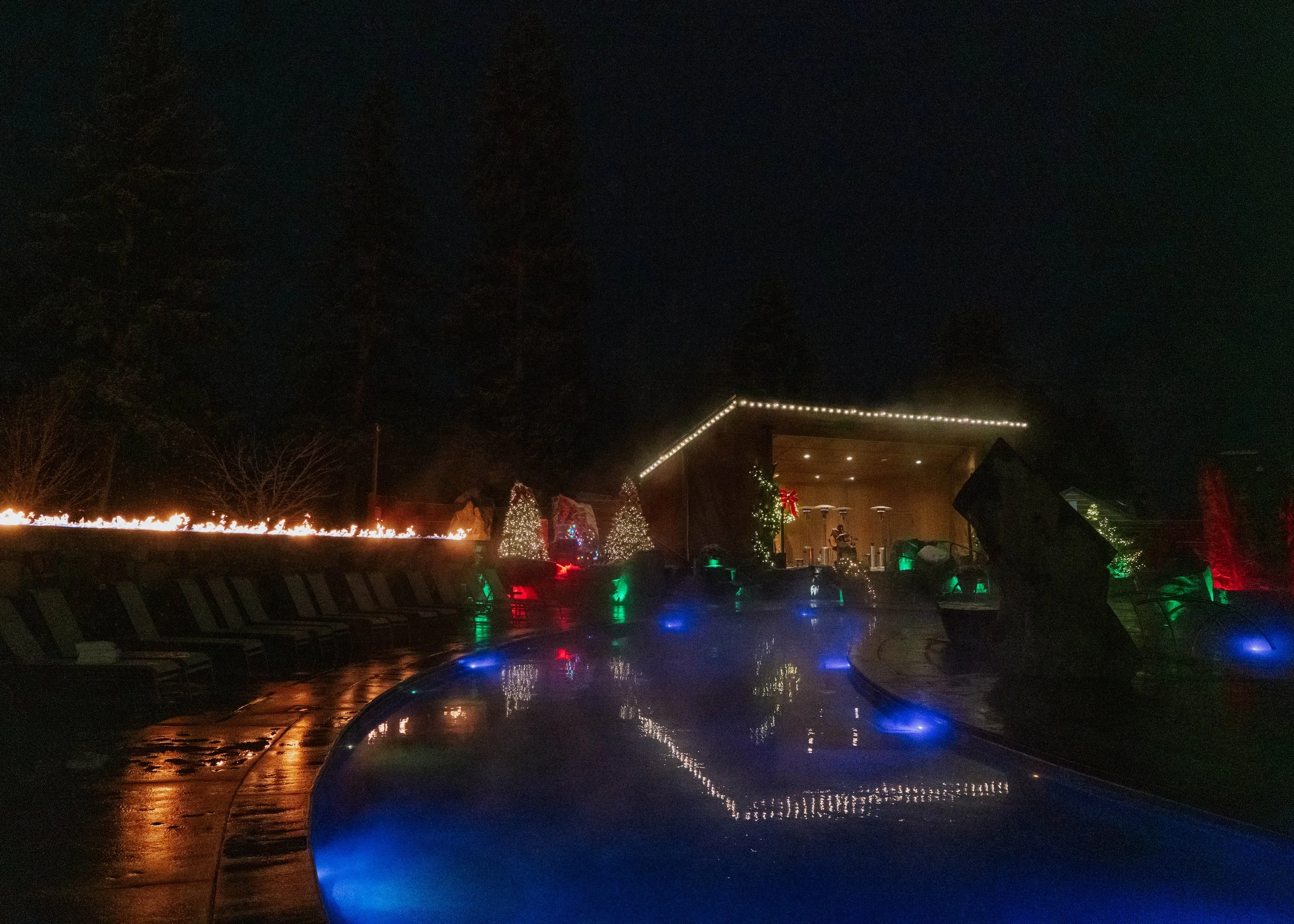 Nighttime scene of a decorated outdoor pool area with holiday lights, Christmas trees, and festive ornaments. The pool reflects colorful lights, and a stage with musical instruments is visible in the background.