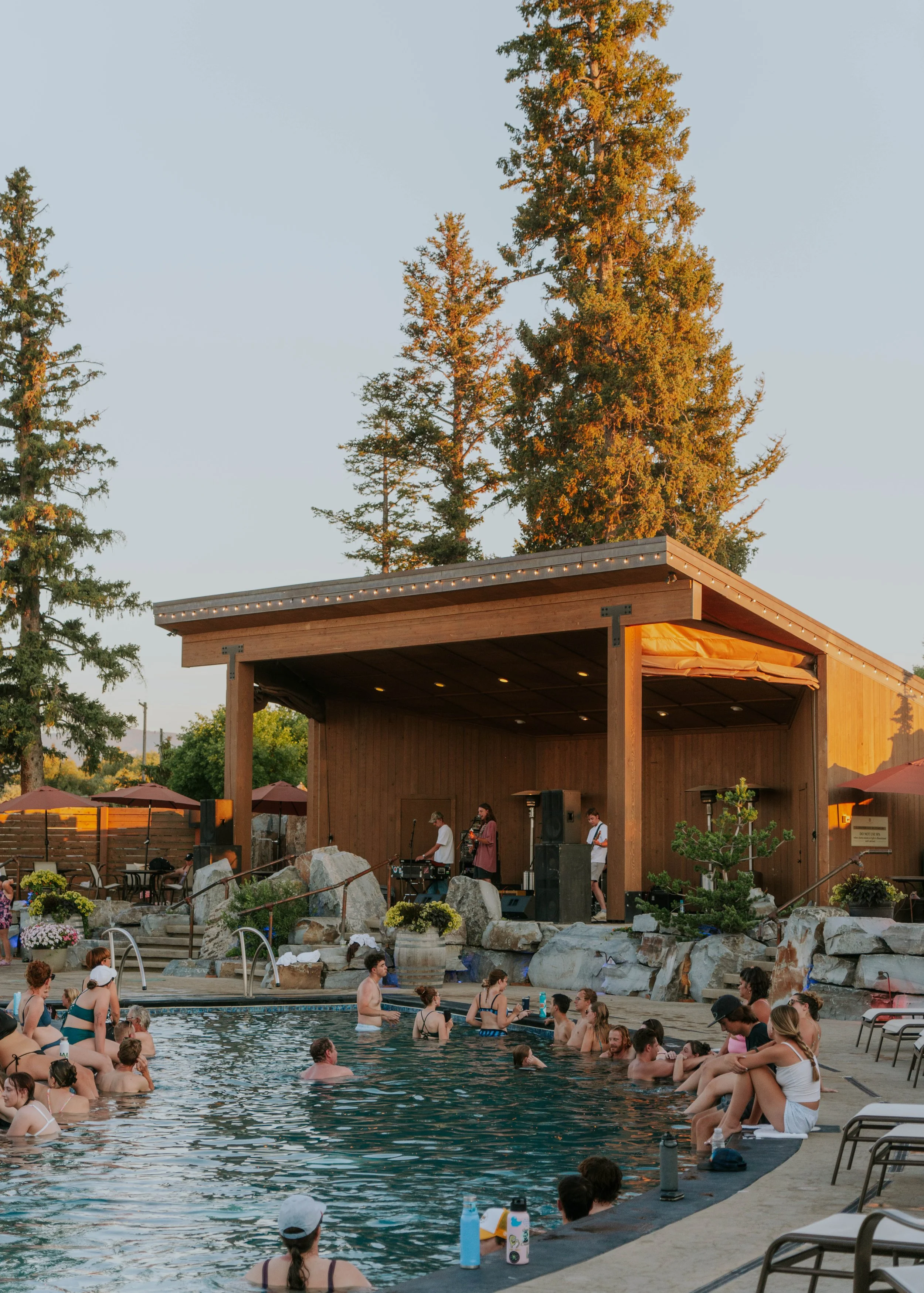 People swimming and relaxing by a pool with a wooden stage and band performing under a covered pavilion, surrounded by trees and lounge chairs.