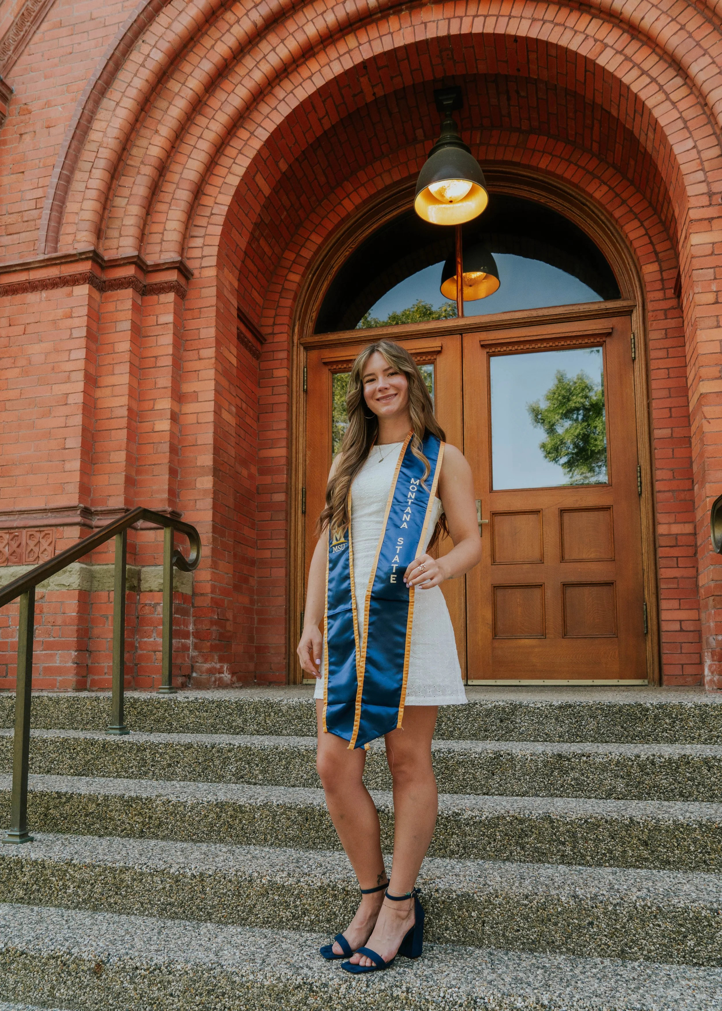 A young woman in a white dress and high heels standing on the steps of a brick building, smiling at the camera. She is wearing a blue stole with gold trim that reads 'Montana State.' The building has a wooden door and an arched brick entrance with a hanging light fixture.