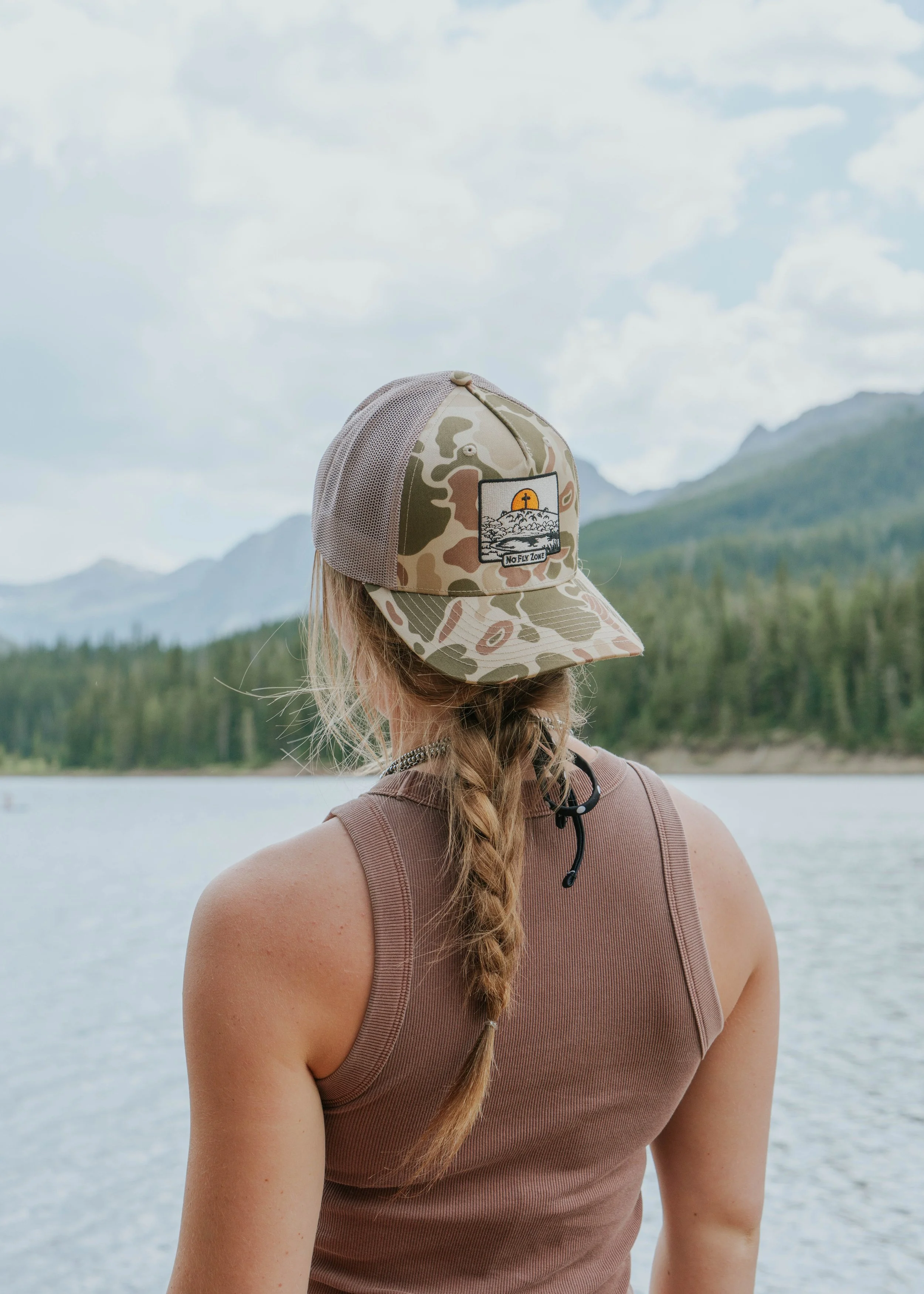 A person with braided hair wearing a camouflage cap and sleeveless brown top standing near a lake with mountains and forest in the background.
