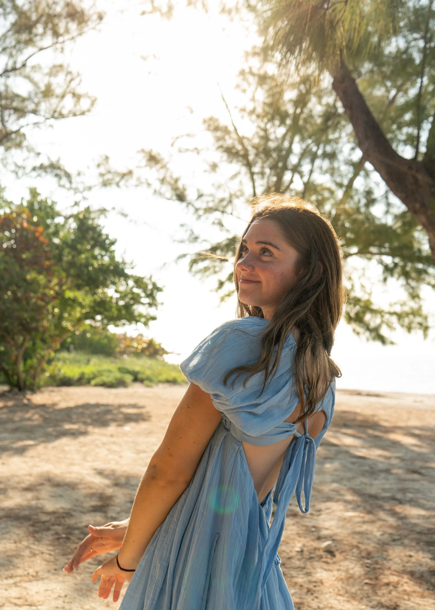 A woman in a blue dress with an open back, smiling and looking to her right, standing outdoors in a sunny, green, tree-filled park or beach area.